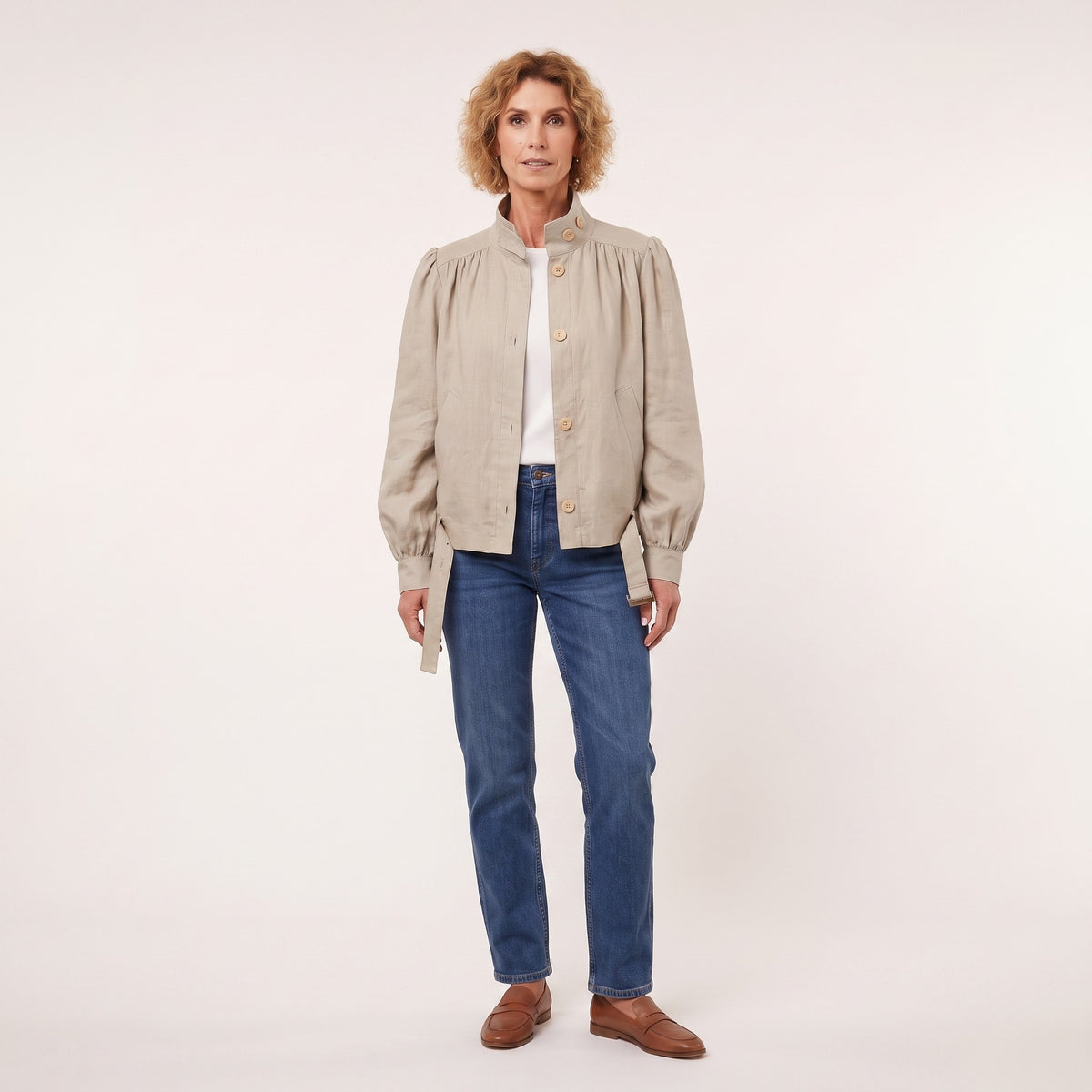 A woman modeling a light beige linen jacket with gathered shoulders and gold buttons, worn over a white top and blue jeans, against a plain off-white studio background.