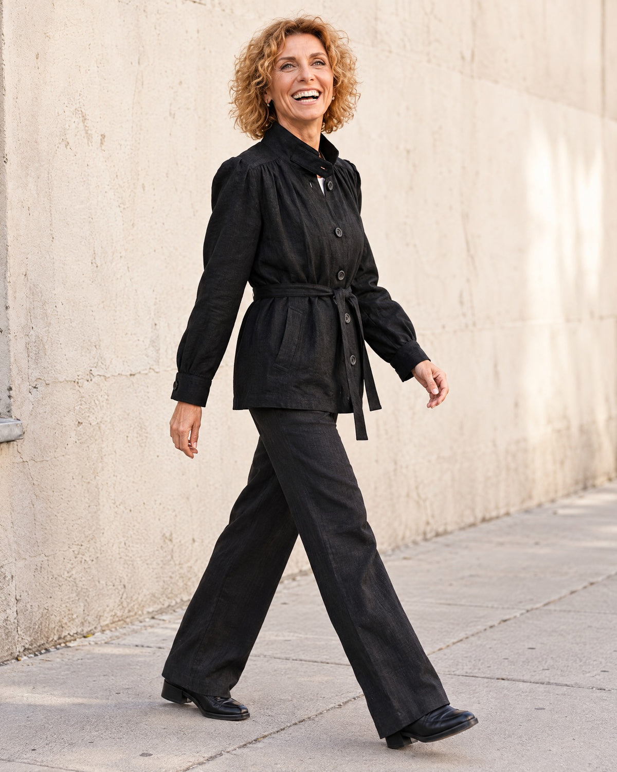 A woman walks in profile near a light concrete wall, wearing a black linen jacket with a stand collar and tie belt, over a white top with dark trousers and dark shoes. #Color_Black