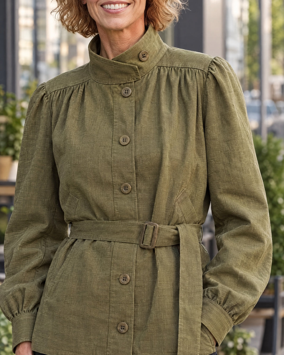 Close-up of an olive green linen jacket showing the stand collar with tab-button closure, wooden buttons, and tie belt with buckle. #Color_Olive green
