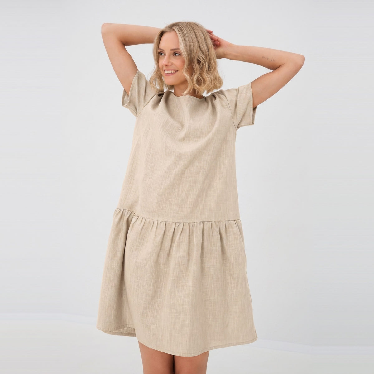 A woman modeling a short-sleeve, tiered linen dress in a light beige color, standing against a plain white studio background.