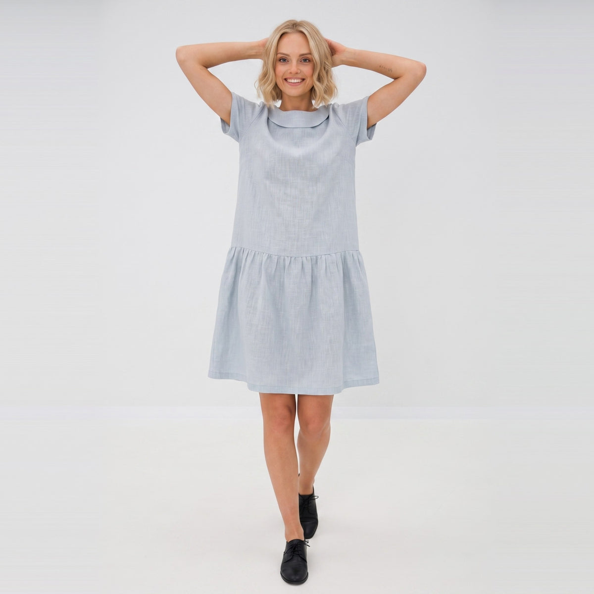 A woman modeling a light blue short-sleeve linen dress with a tiered skirt, standing against a plain white studio background.