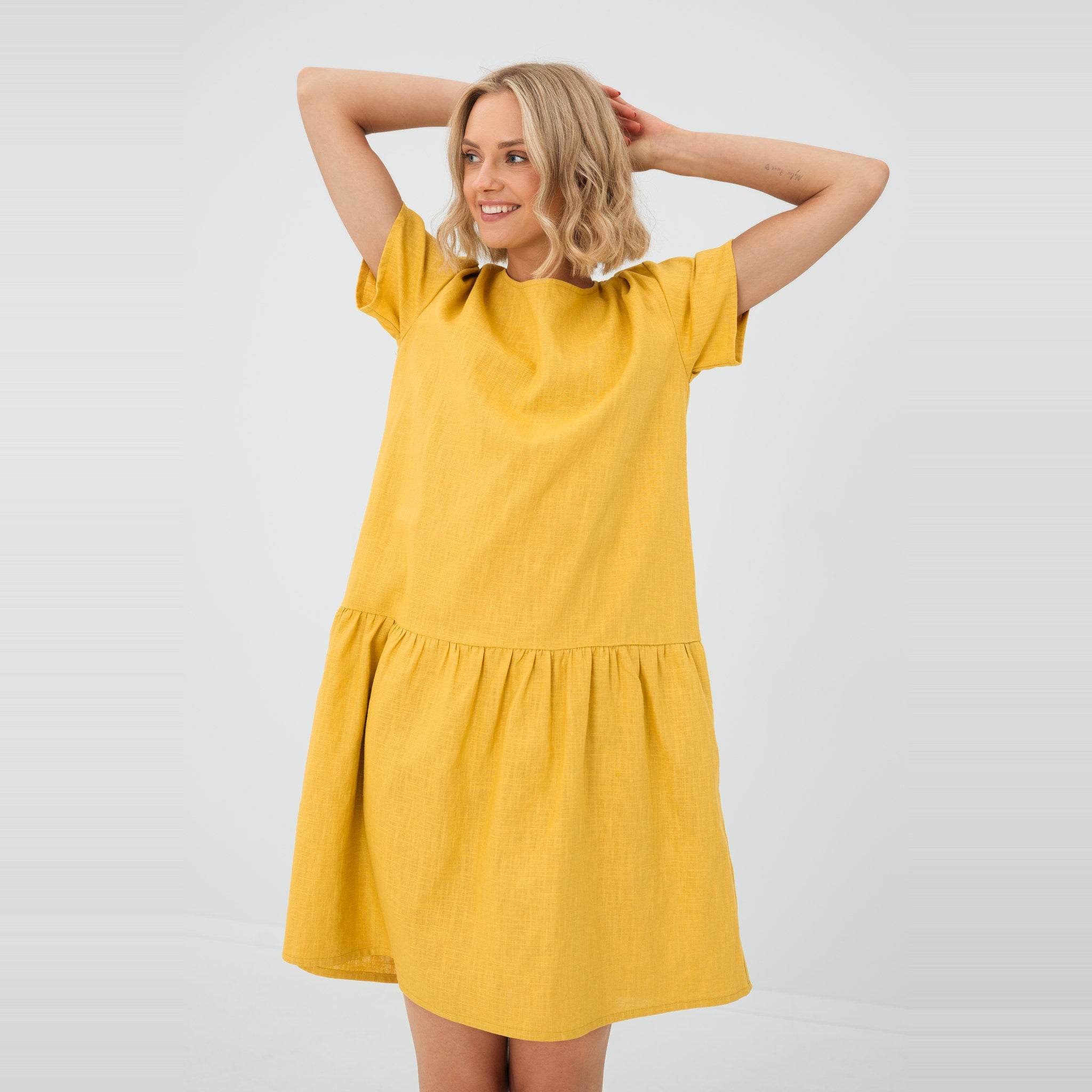 A woman modeling a short-sleeve tiered linen dress in a warm mustard yellow color, set against a plain white studio background.