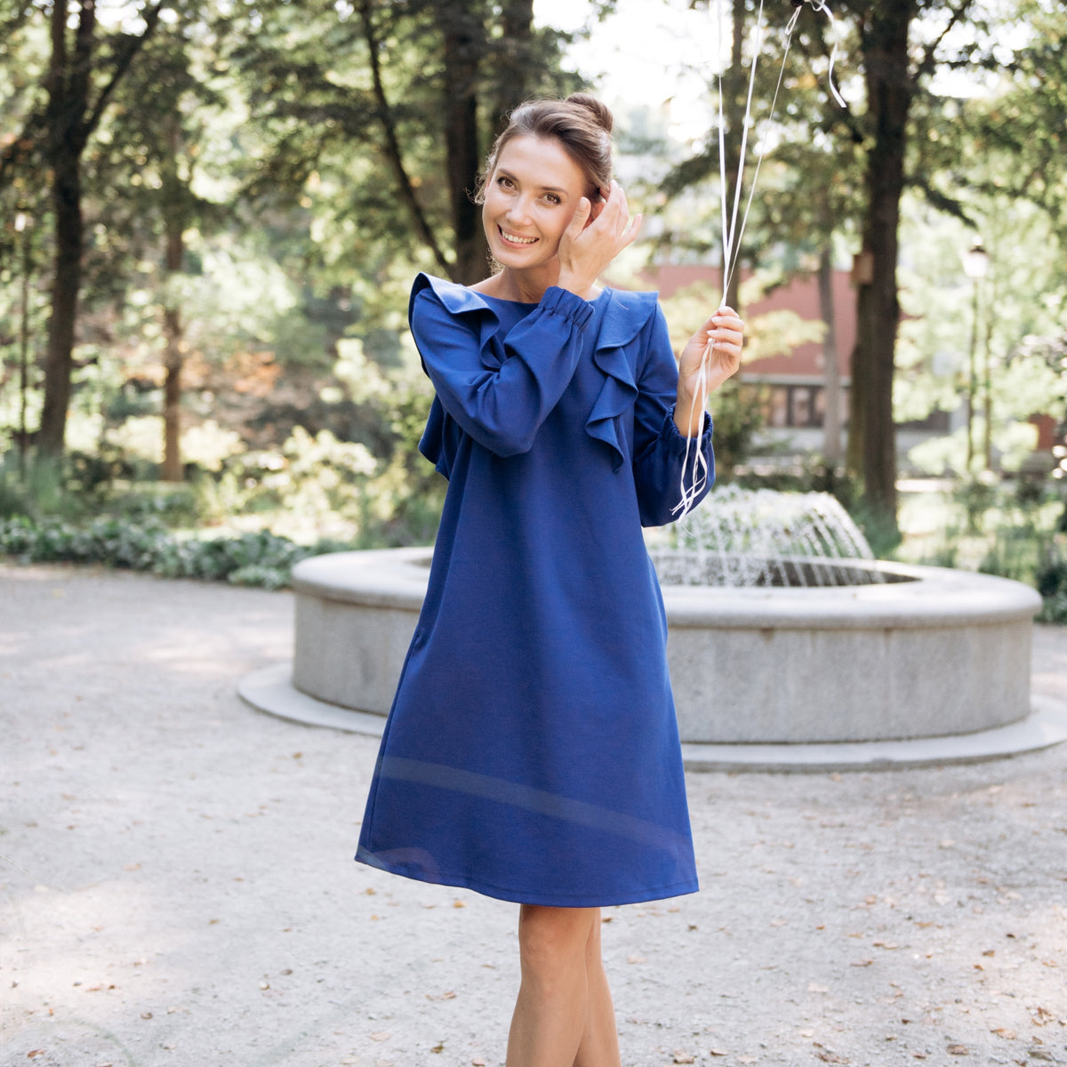 A woman wearing a royal blue midi dress with ruffled long sleeves stands in a park in front of a stone fountain, holding a decorative silver wand.