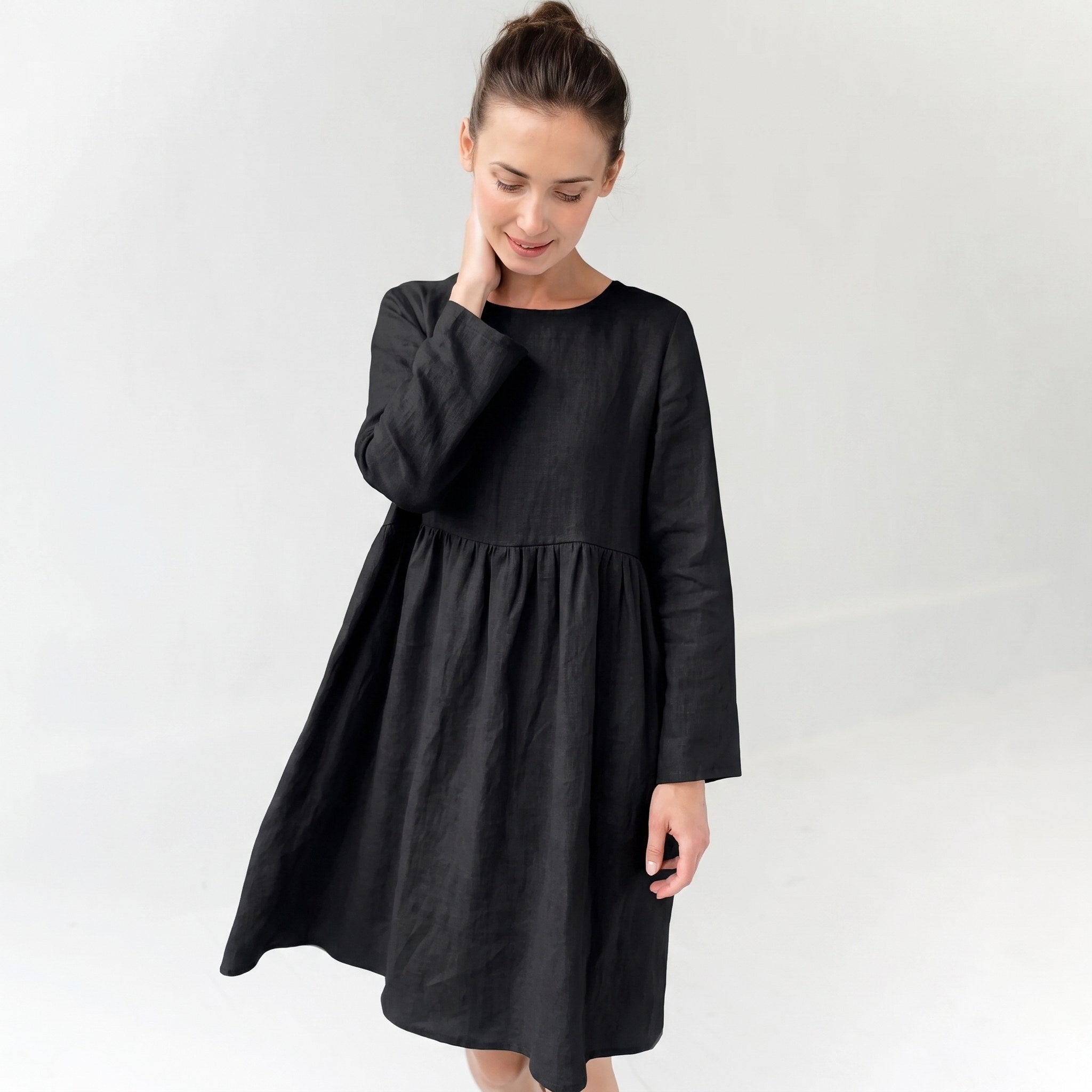 A woman wearing a black long-sleeve linen dress with a gathered waist, standing against a plain white studio background.