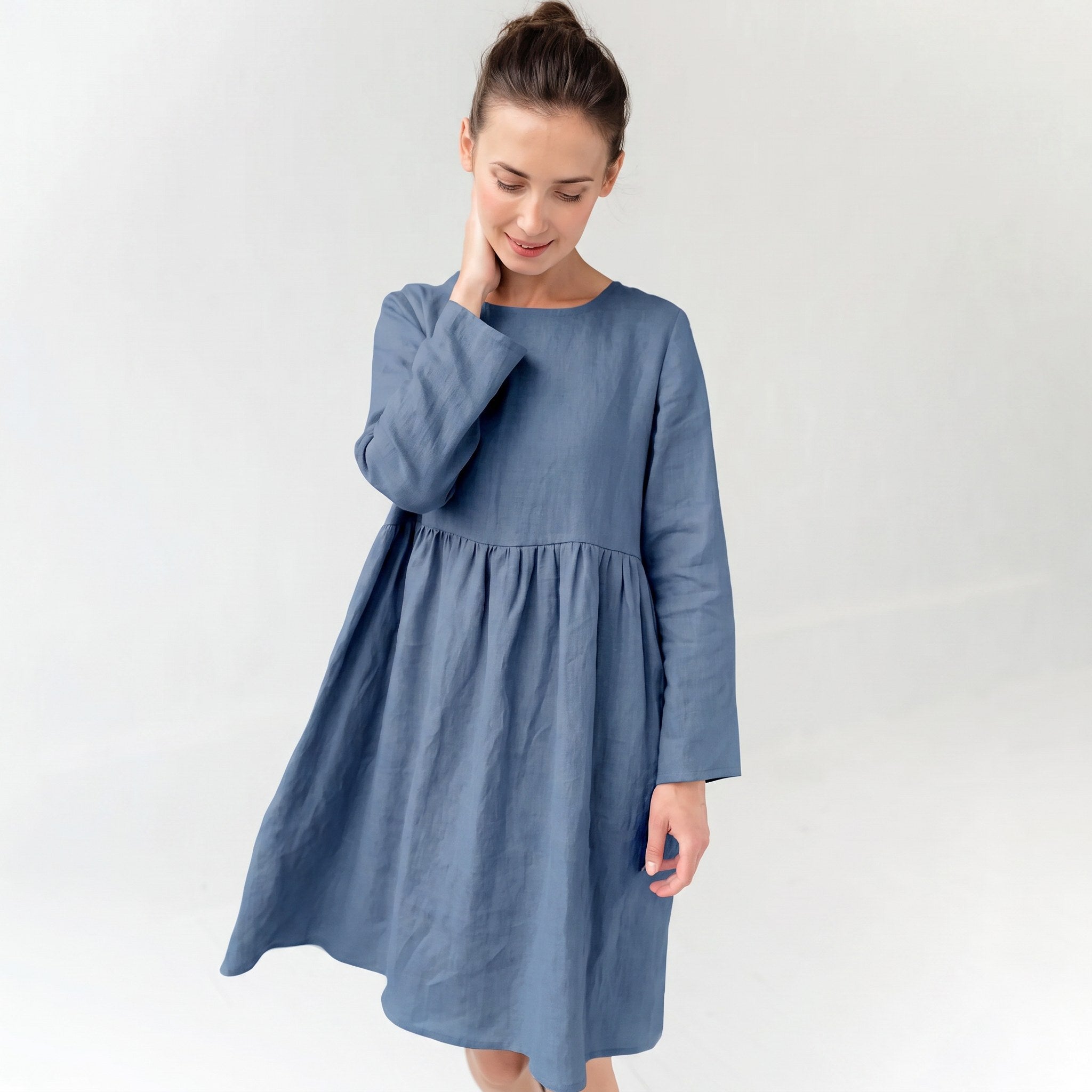 A woman wearing a dusty blue long-sleeve linen dress with a gathered waist, standing against a plain white studio background.
