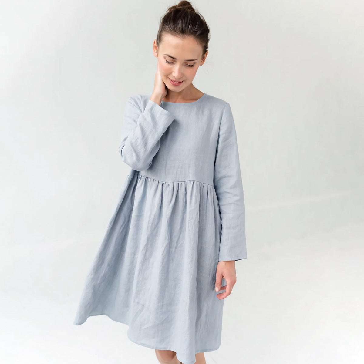 A woman wearing a light blue long-sleeve linen dress with a gathered waist, standing against a plain white studio background.