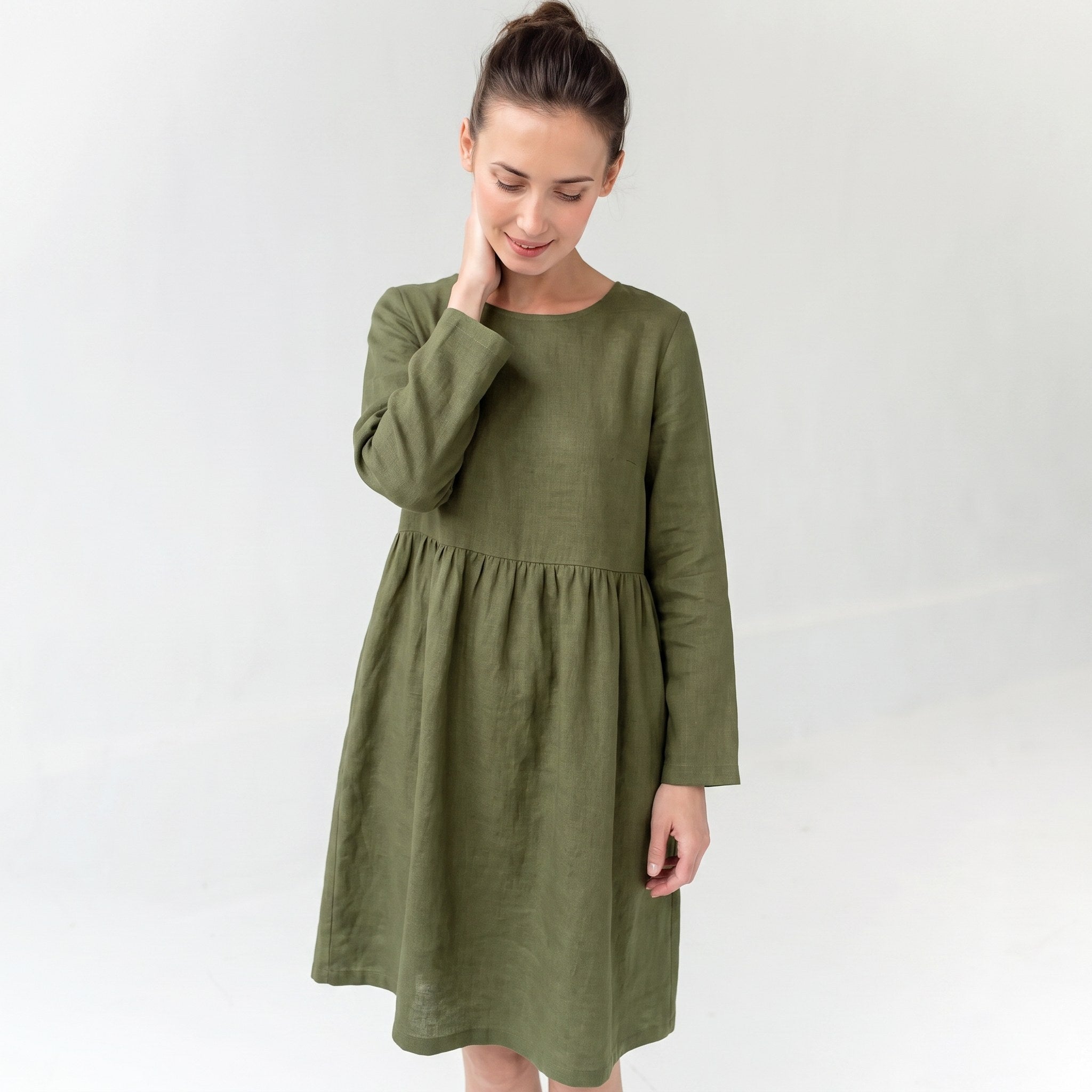 A woman wearing a long-sleeved olive green linen dress with a gathered waist, standing against a plain light gray background.