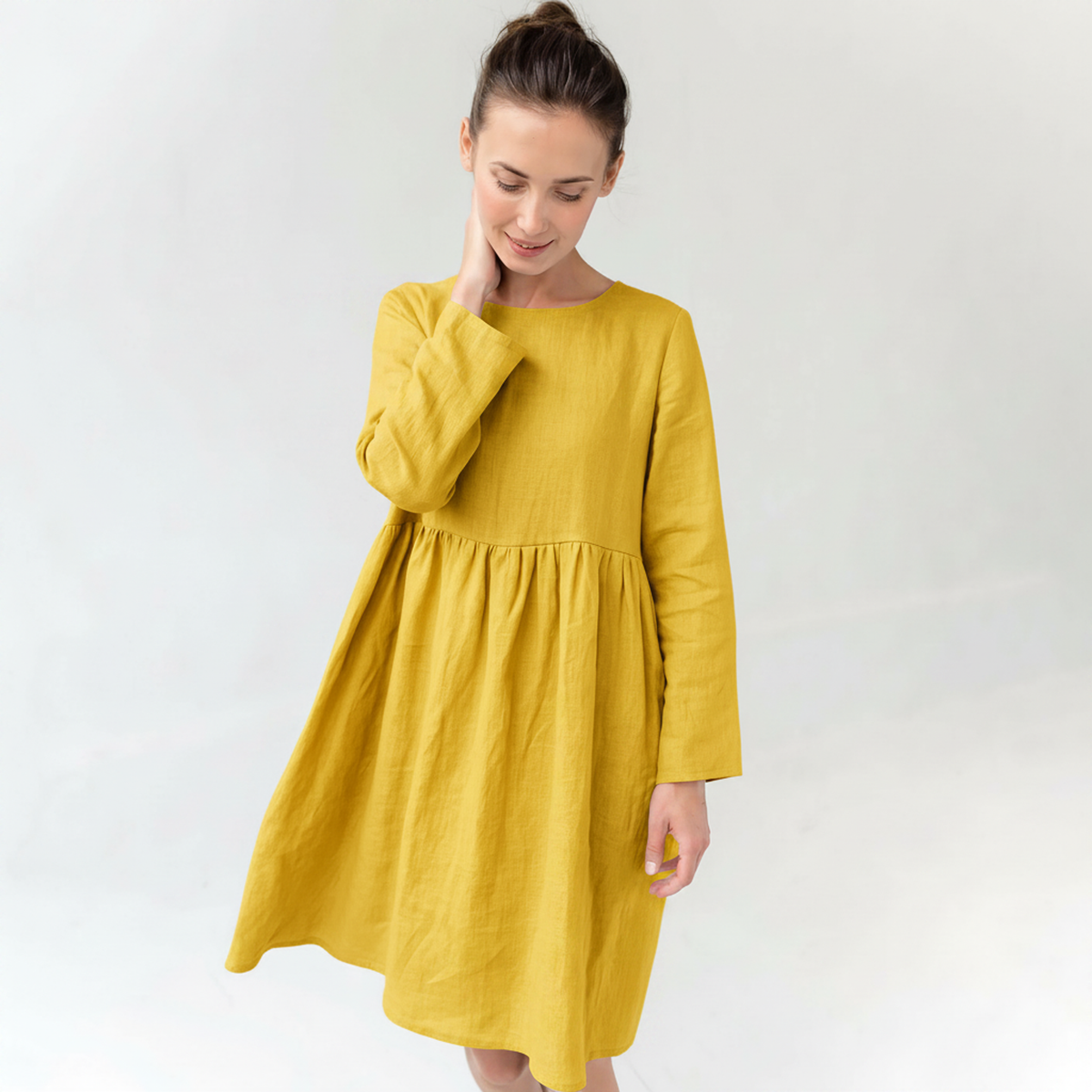 A woman wearing a mustard yellow linen dress with long sleeves and a gathered waist, standing against a plain white studio background.