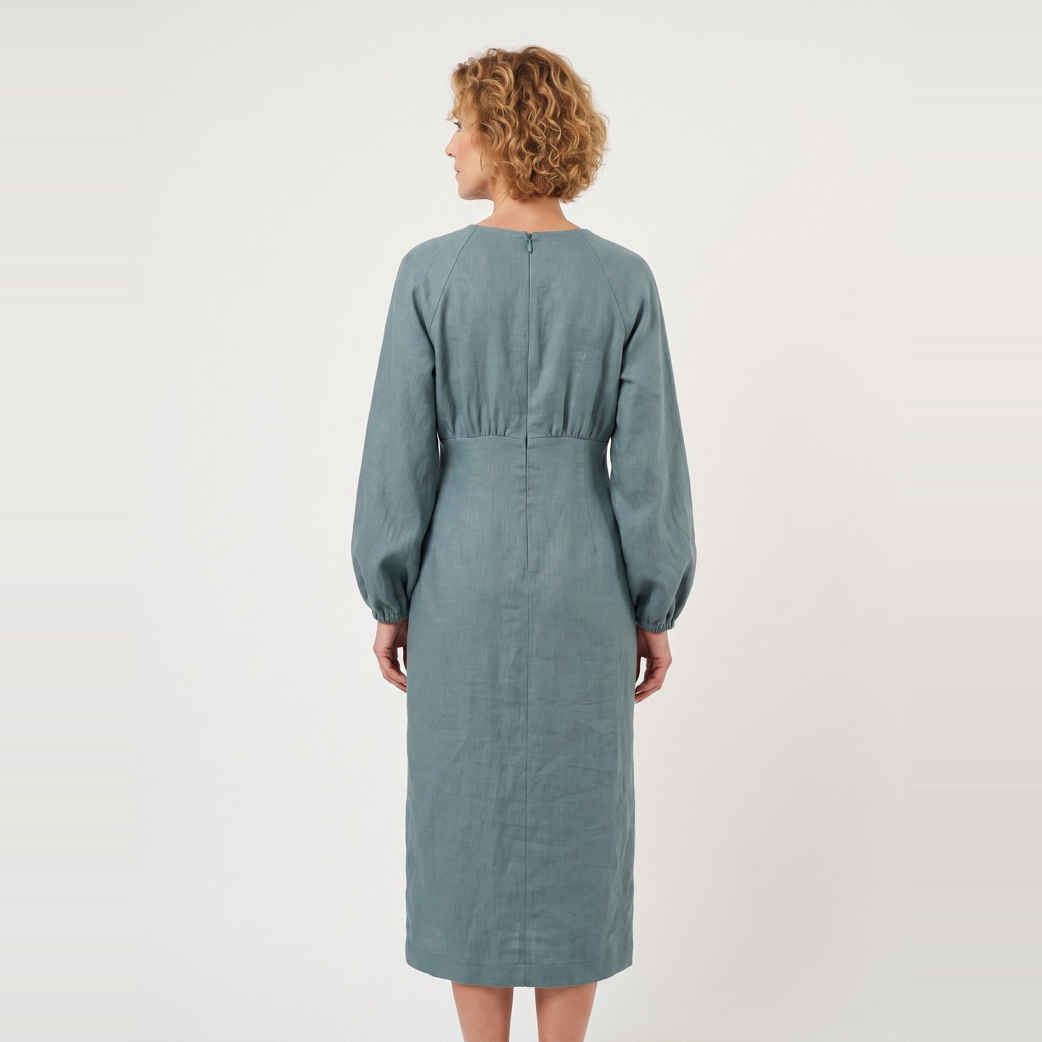 A woman viewed from the back wearing a long-sleeve midi dress in a muted dusty blue-green color, set against a plain off-white studio background.