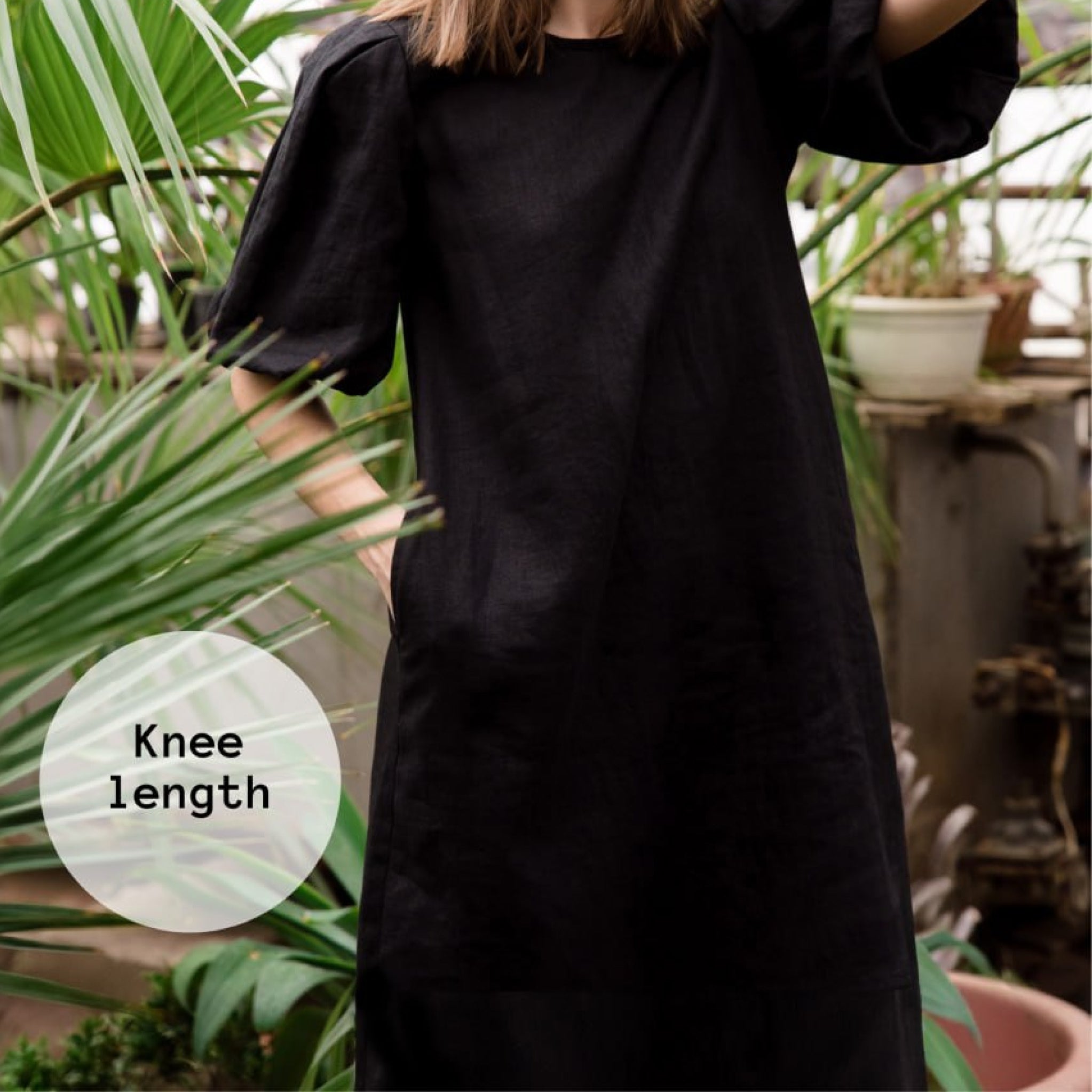 A person wearing a loose black knee-length dress or tunic with short puff sleeves, standing among green plants. A translucent circular label on the left reads 'Knee length'.