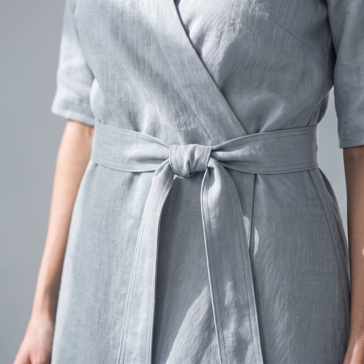A close-up of a person wearing a light gray wrap-style dress with a matching waist tie, shown against a plain gray background.