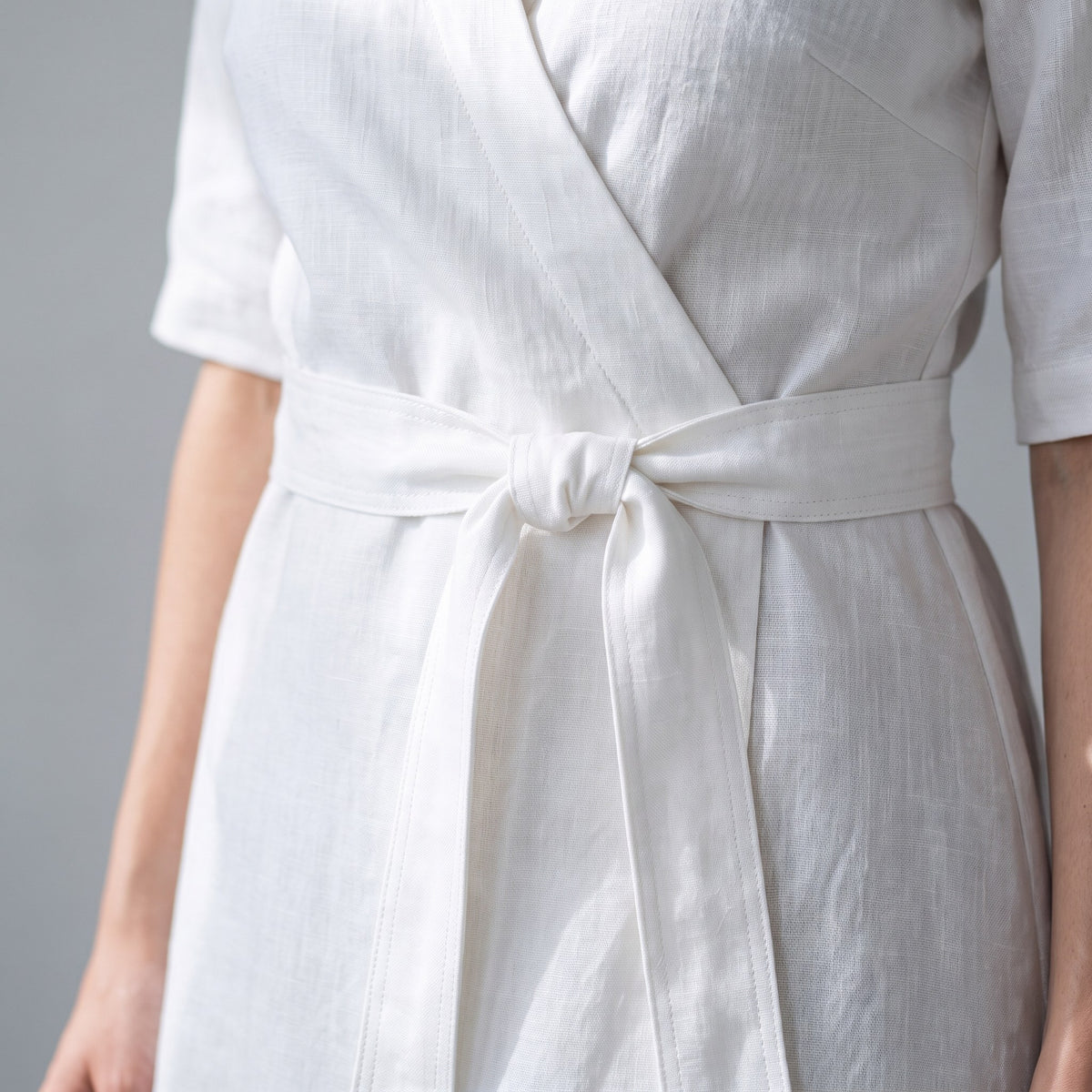 A close-up of a person wearing a white linen wrap dress with a matching tie belt, showing the textured fabric and waist detail against a neutral gray background.