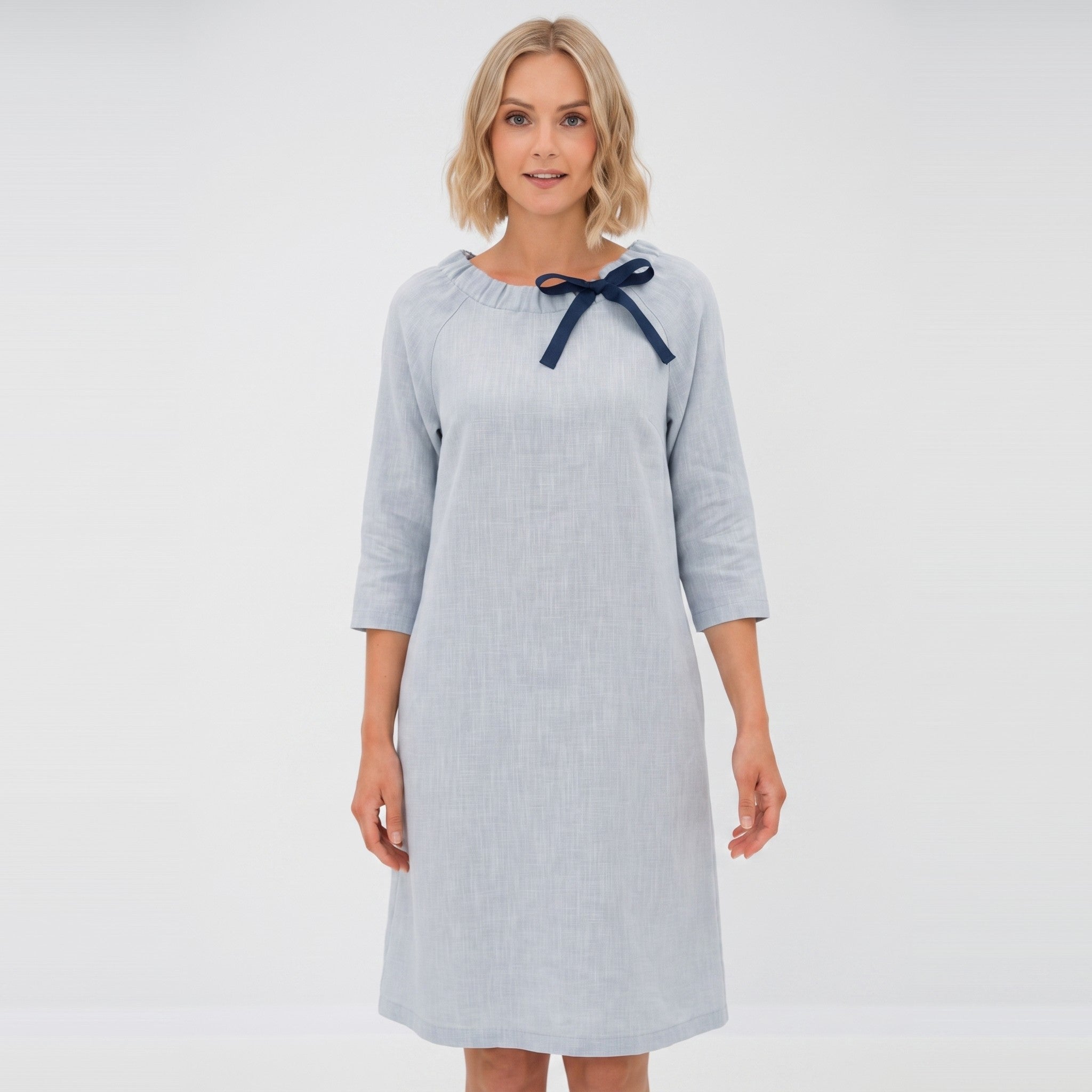 A woman modeling a light blue long-sleeve dress with a navy blue bow detail at the neckline, set against a plain white background.