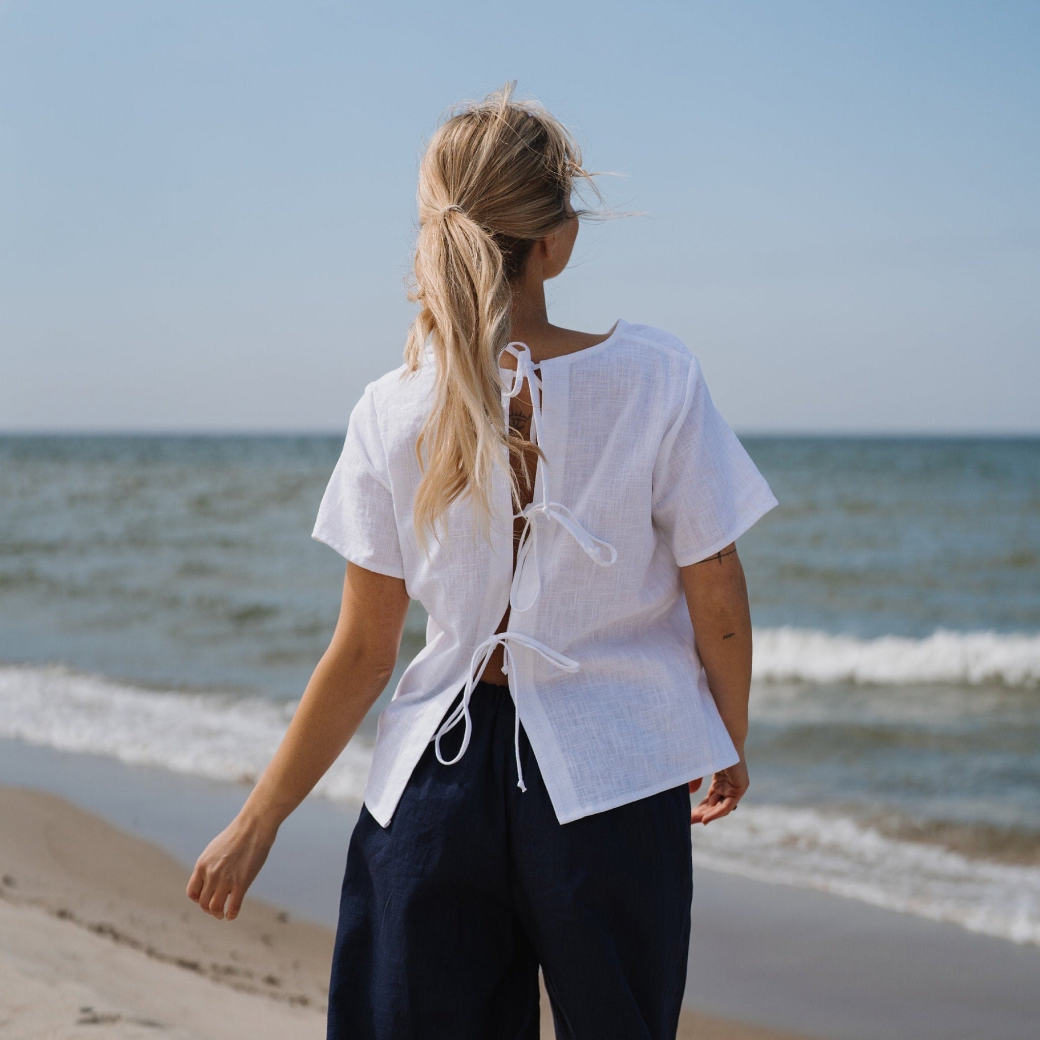 A woman stands on a sandy beach facing the ocean, wearing a white short-sleeve linen blouse with lace-up back details and dark navy pants.
