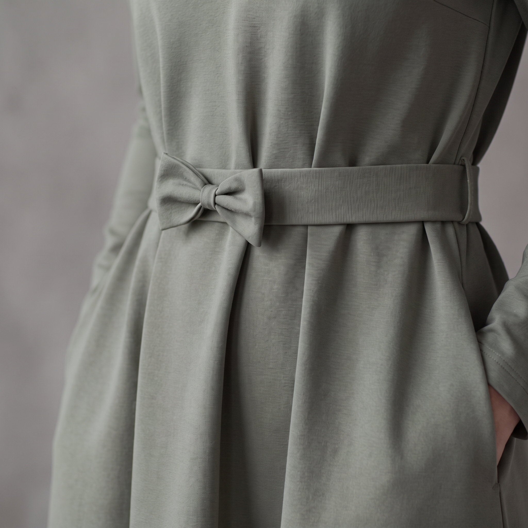 A sage green dress with a matching fabric tie belt at the waist, showing the draped skirt and side pocket detail.