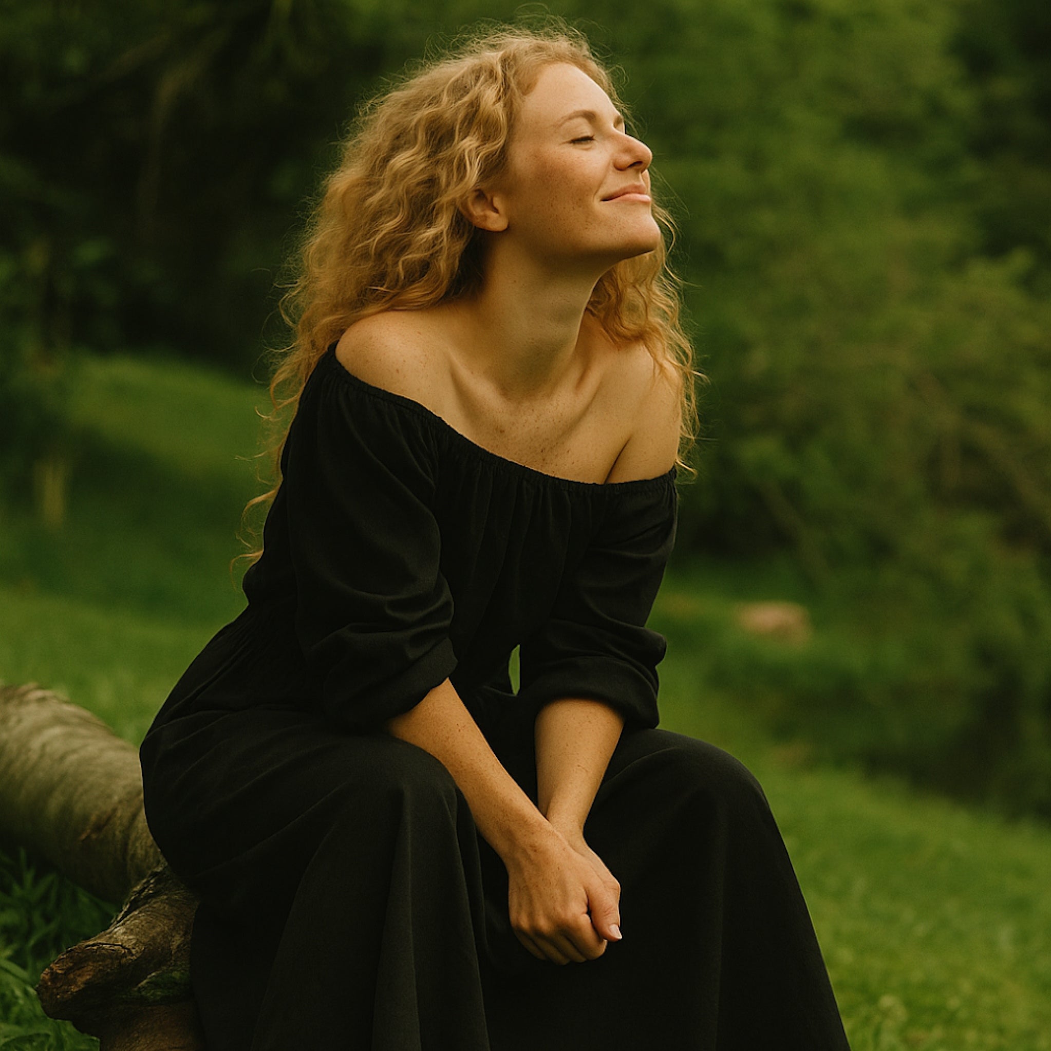 A woman with curly blonde hair sits outdoors on a log in a grassy, wooded setting, wearing a black off-the-shoulder long-sleeve dress.