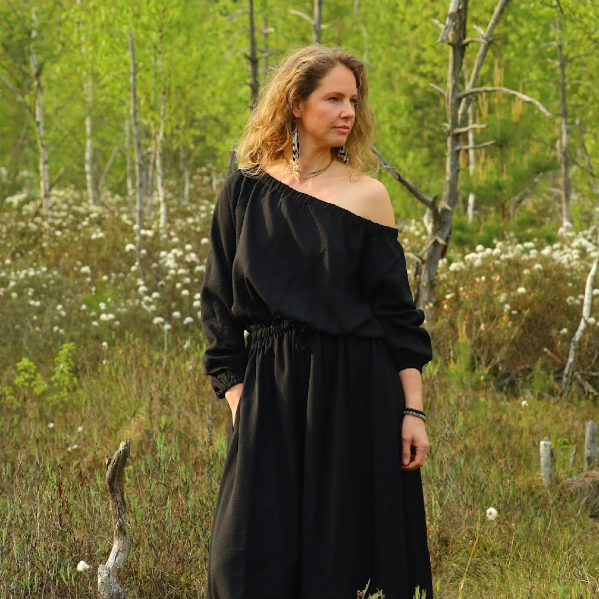 A woman wearing a black off-the-shoulder maxi dress stands in a grassy, wooded outdoor setting with birch trees and small white flowers.