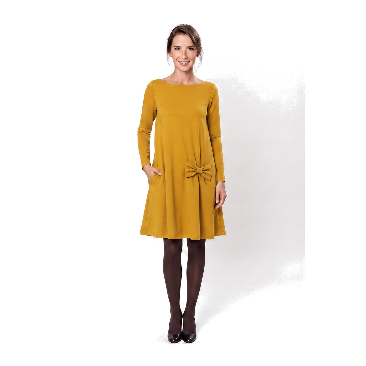 A woman modeling a mustand yellow long-sleeve A-line dress with a boat neckline, side pockets, and a bow detail at the waist.