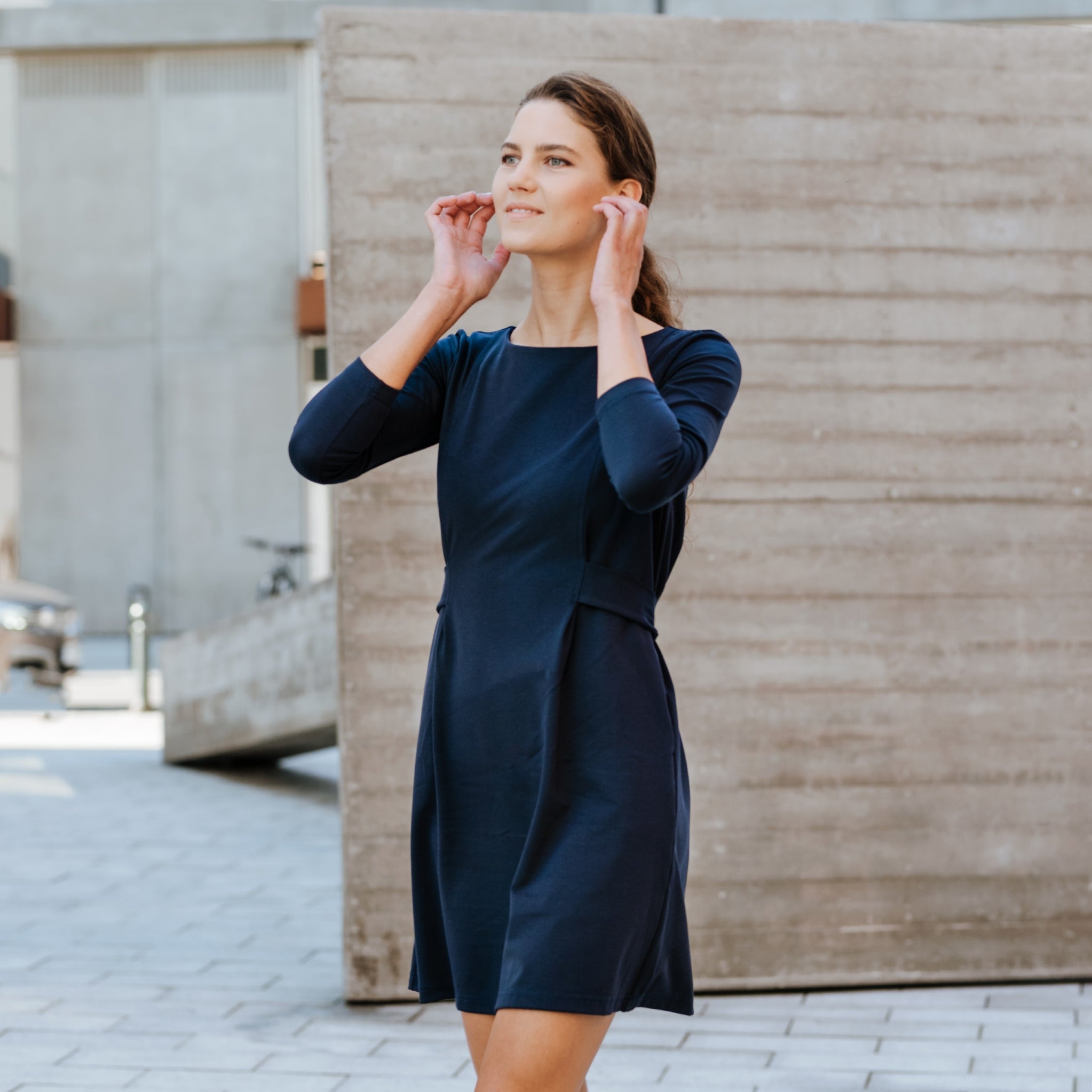 A woman wearing a dark navy blue knee-length dress with three-quarter sleeves, standing outdoors in front of a concrete wall.