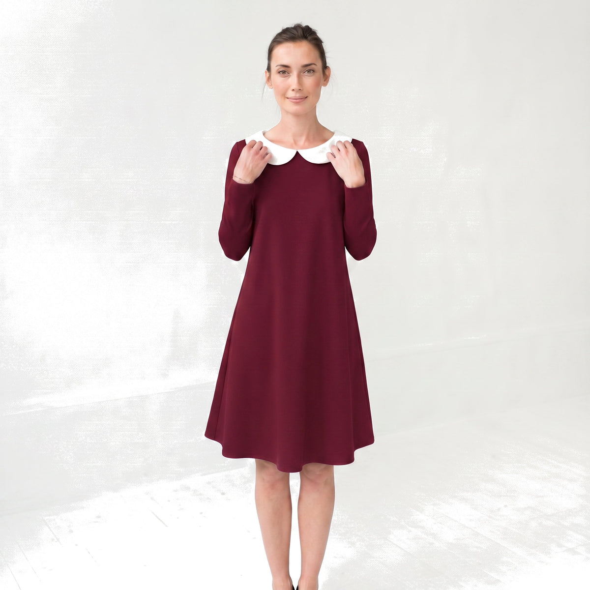 A woman modeling a long-sleeve A-line dress in deep burgundy with a contrasting white Peter Pan collar, standing against a light gray textured studio background.