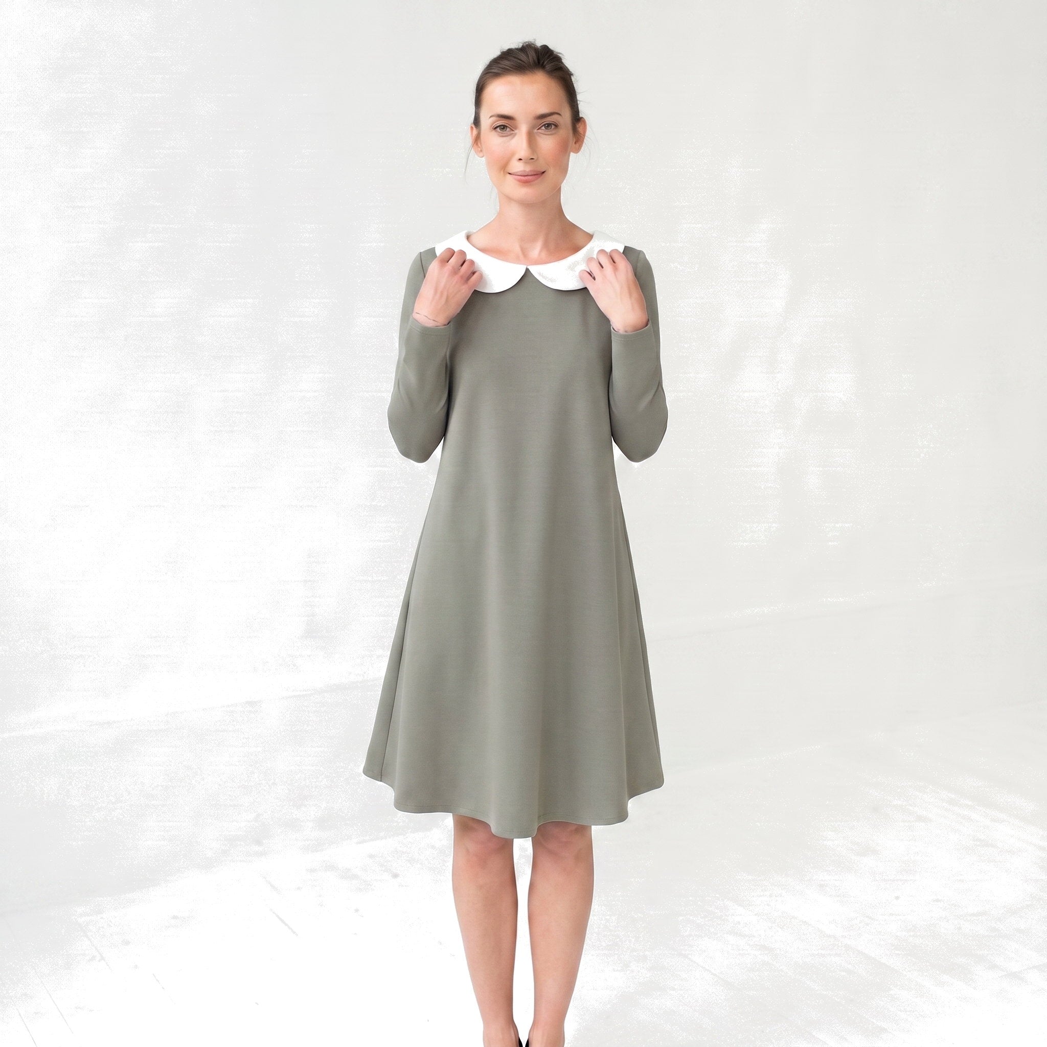 A woman modeling a long-sleeve knee-length dress in muted sage green with a contrasting white Peter Pan collar, standing against a light gray textured studio backdrop.