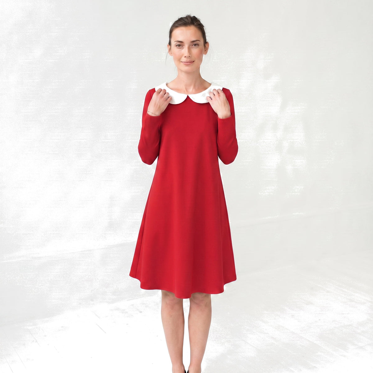 A woman modeling a red long-sleeve A-line dress with a contrasting white Peter Pan collar, standing against a light gray textured studio backdrop.