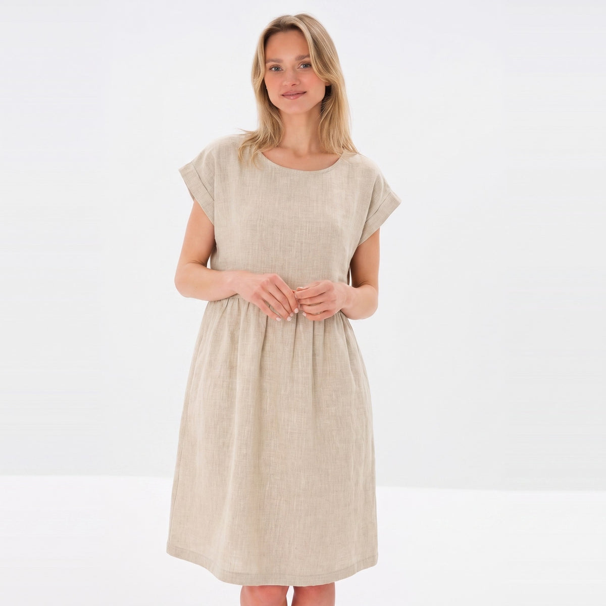 A woman modeling a light beige linen dress with short cap sleeves and a gathered waist, set against a plain white studio background.