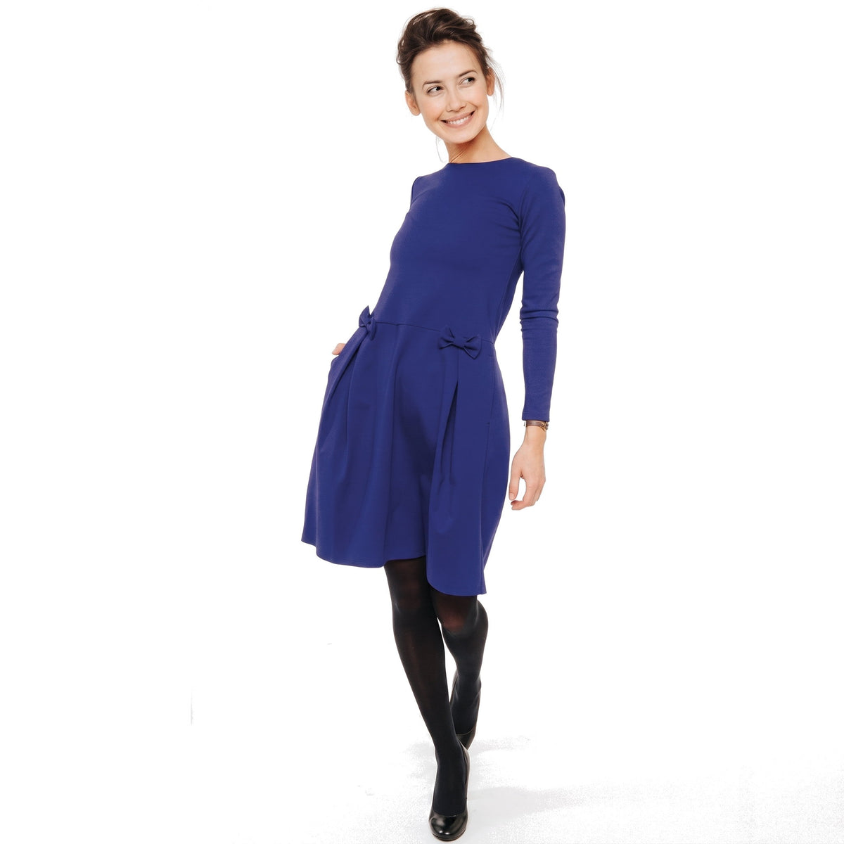 A woman wearing a royal blue long-sleeve knee-length dress with a structured waist and bow details, paired with black tights and black shoes.