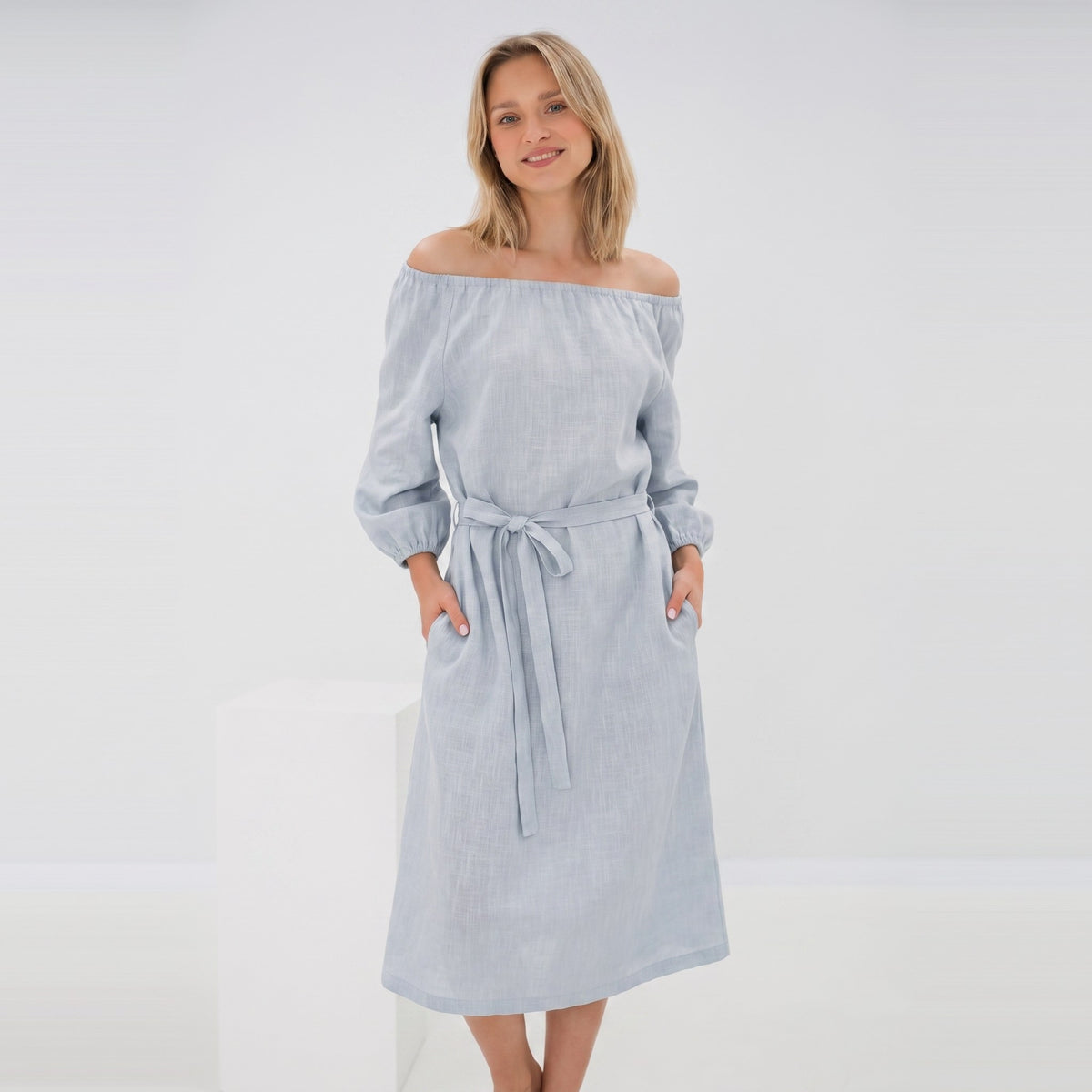 A woman modeling a light blue off-the-shoulder midi dress with a tie waist belt and front pockets, set against a clean white studio background.