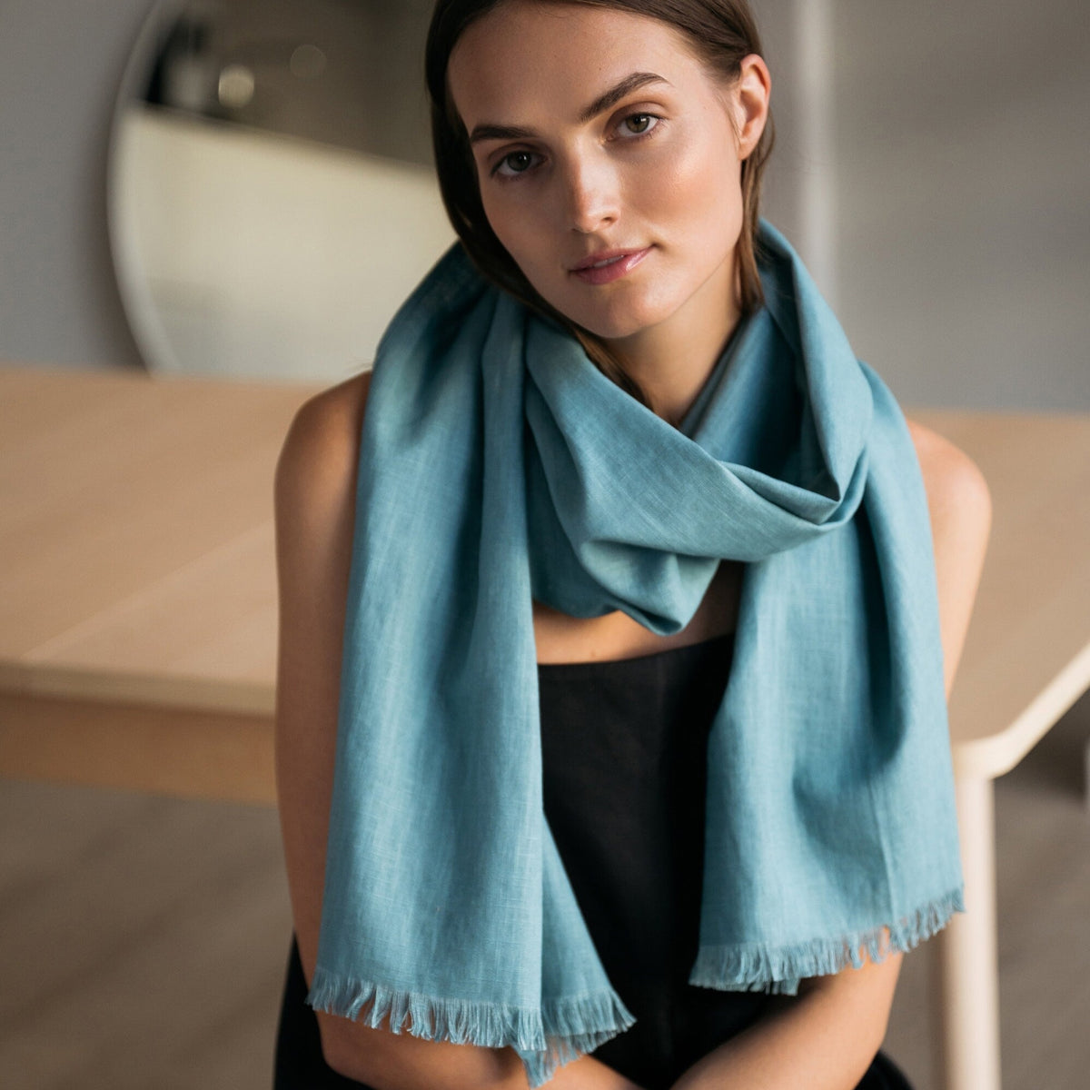 A woman wearing a teal fringed scarf draped over her shoulders, paired with a black top, in a softly lit indoor setting.