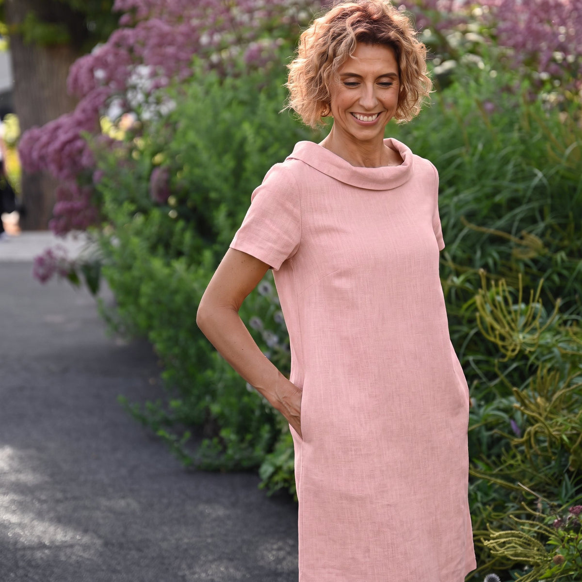 A close-up lifestyle shot of a woman with curly light brown hair wearing a light pink short-sleeve dress with a wide collar and bow detail on the back.