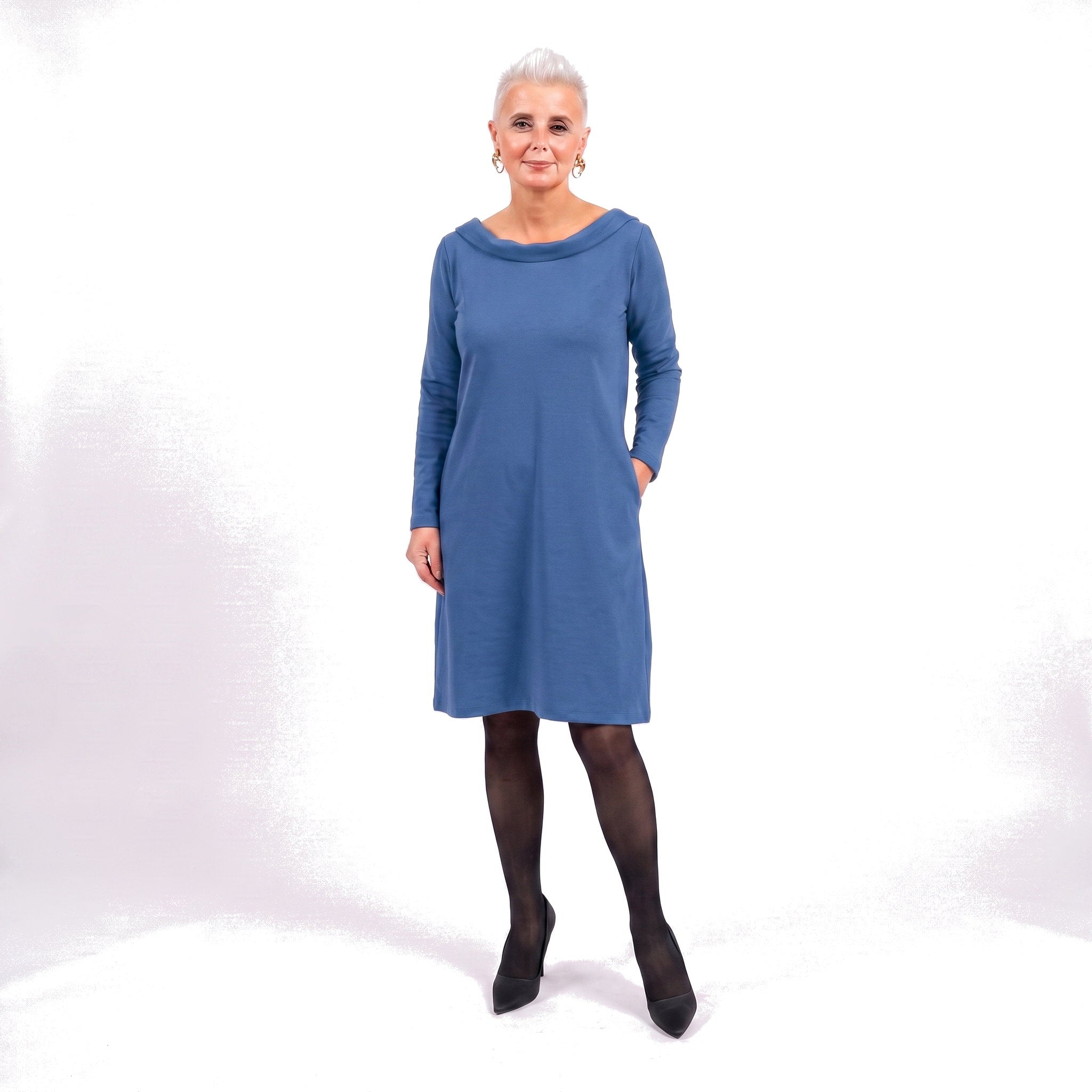 An older woman with short gray hair stands against a white studio background wearing a long-sleeve blue shift dress, black tights, and black pointed shoes.