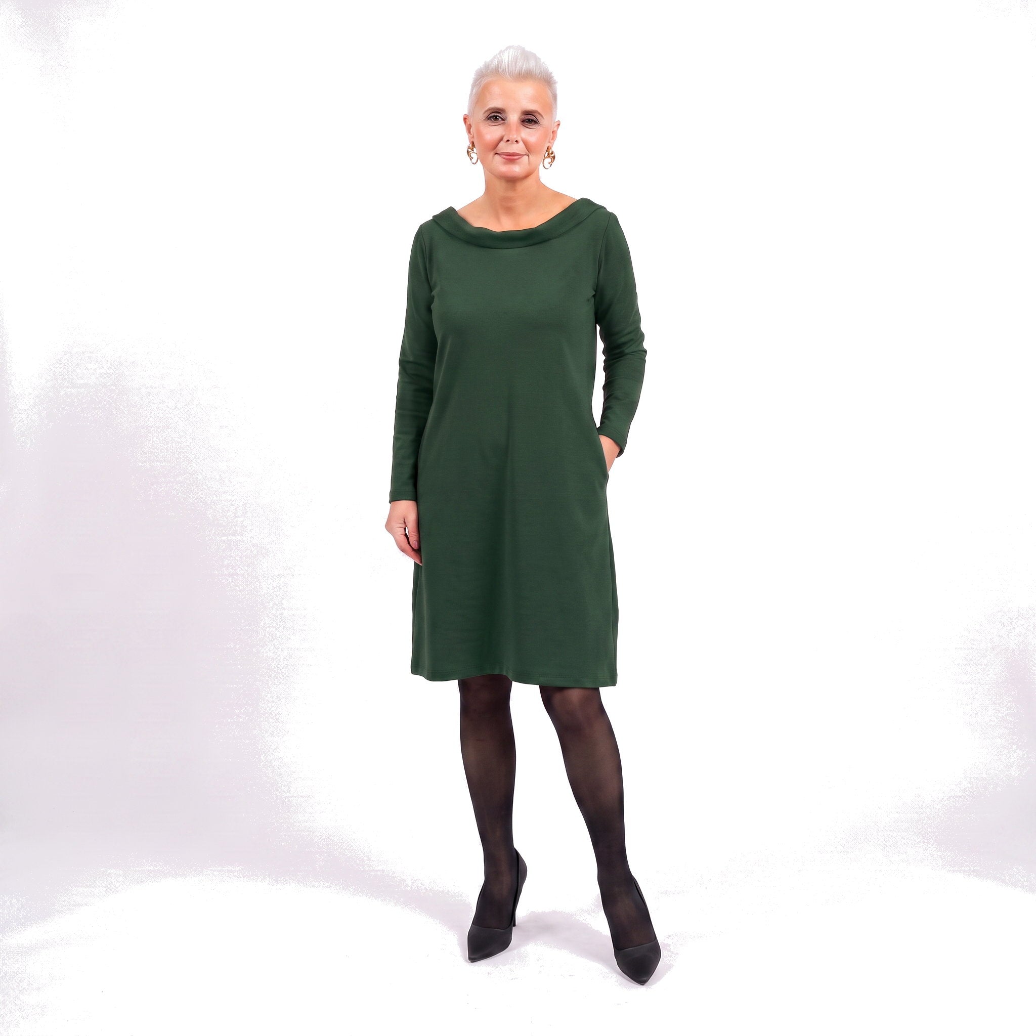 An older woman with short gray hair models a dark green long-sleeve shift dress with pockets, paired with black tights and black pointed-toe heels, against a white studio background.