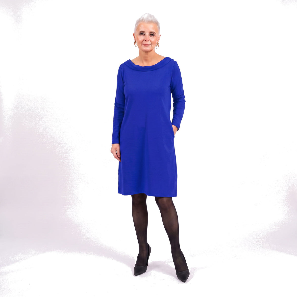A woman modeling a bright royal blue long-sleeve shift dress with side pockets, paired with black tights and black pointed-toe heels, against a white studio background.