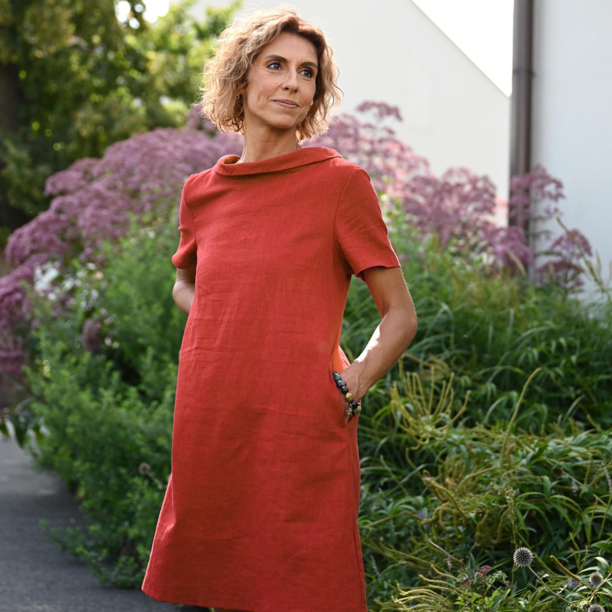 A woman wearing a rust-red linen dress with short sleeves and a cowl neck, standing outdoors in a garden setting with green plants and purple flowers.