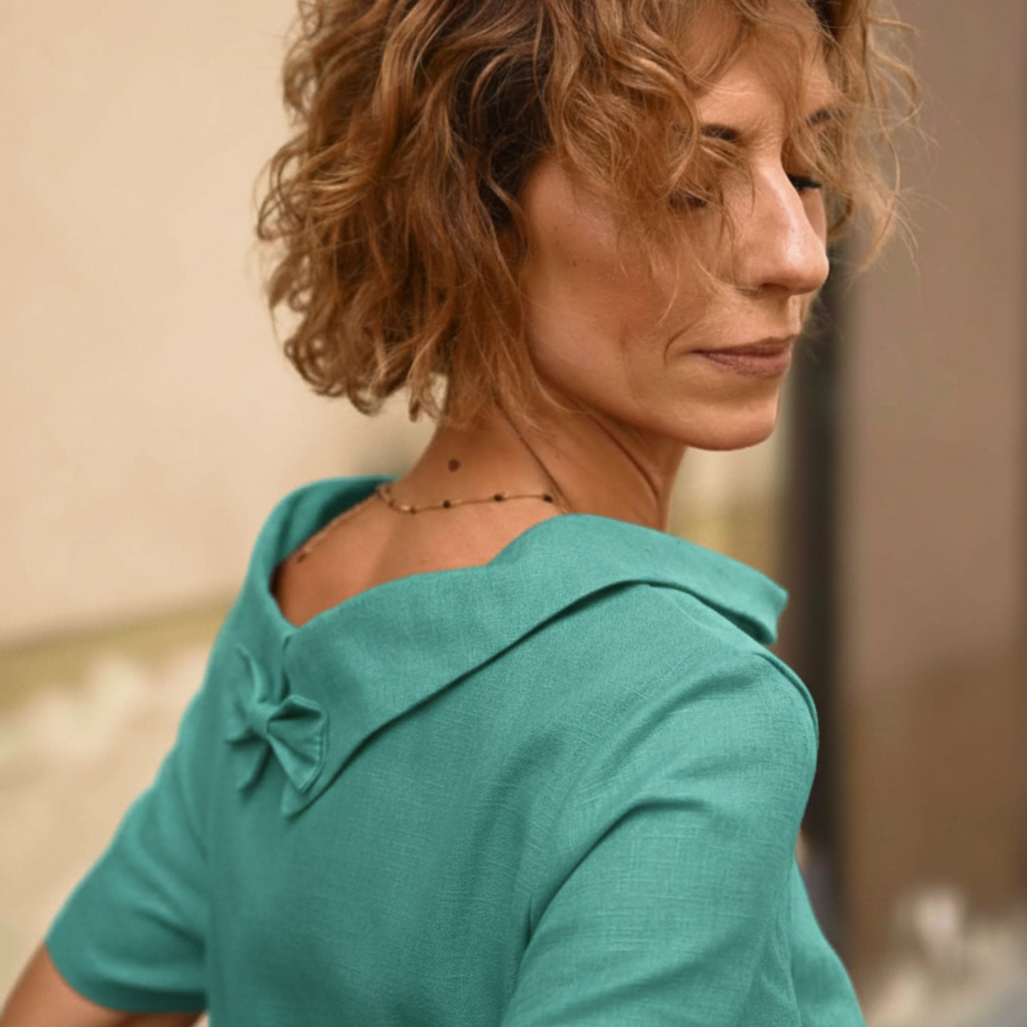 A close-up lifestyle shot of a woman with curly light brown hair wearing a teal green short-sleeve dress with a wide collar and bow detail on the back.
