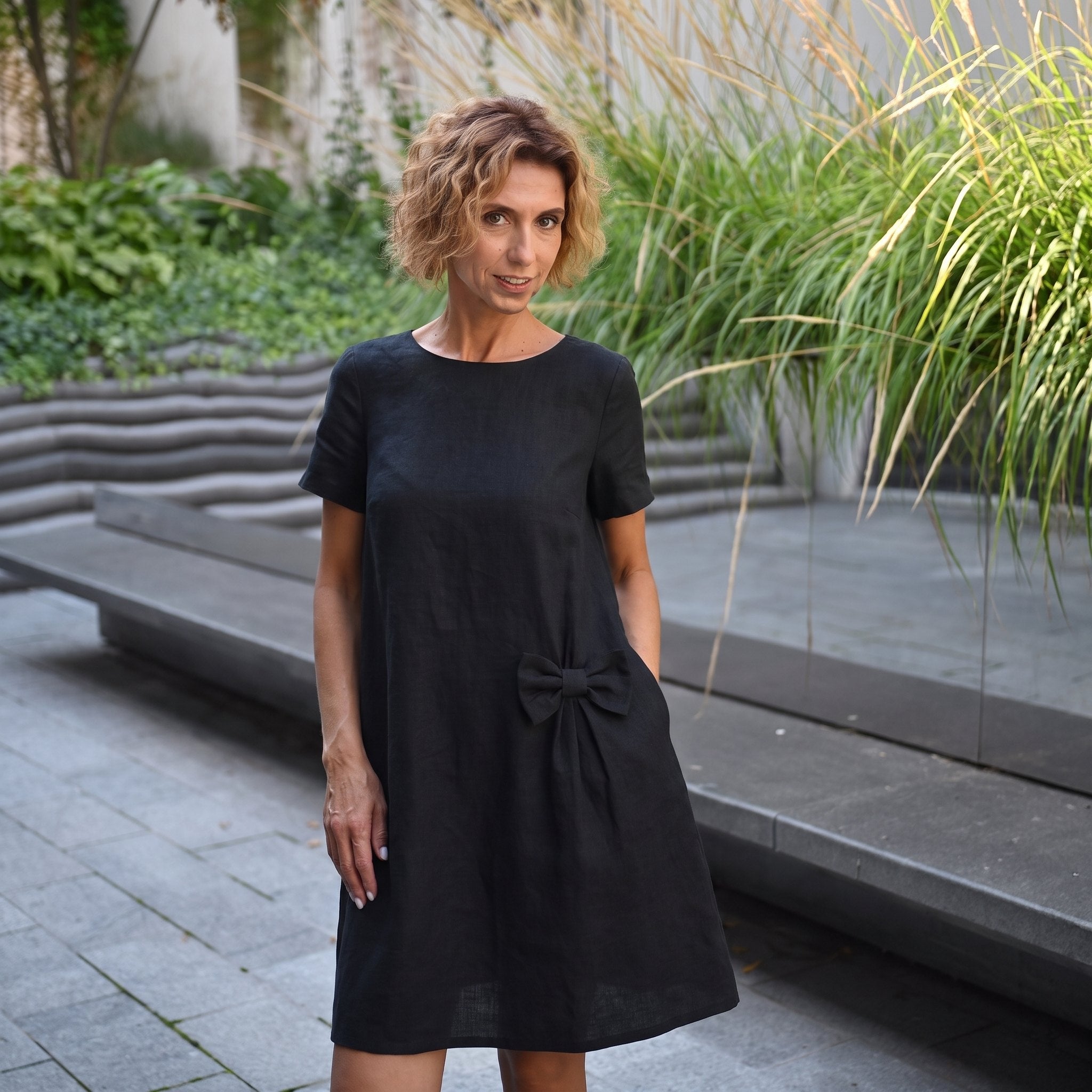 A woman wearing a black short-sleeve linen dress with a bow detail at the waist, standing in an outdoor garden setting with stone steps and green plants.