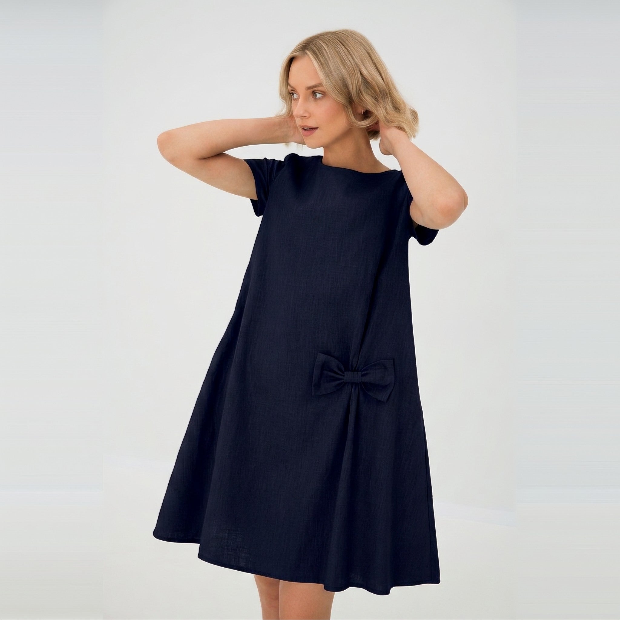 A woman modeling a short-sleeve navy blue linen dress with a bow detail on the front, set against a plain light gray background.