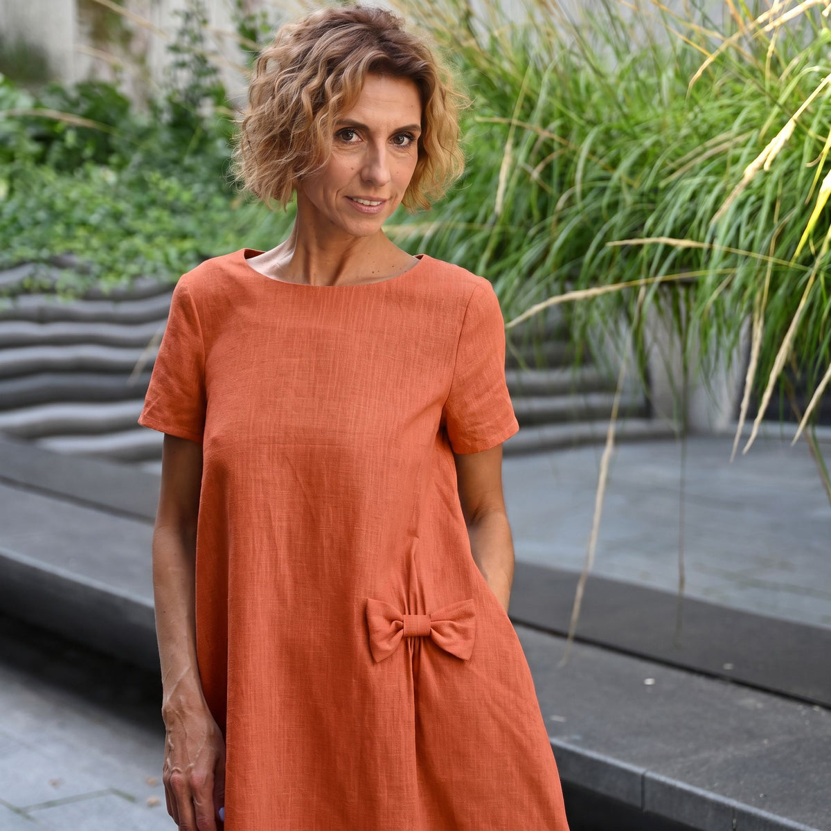 A woman wearing a short-sleeved terracotta orange linen dress with a gathered bow detail at the waist, standing outdoors in front of modern concrete steps and green plants.