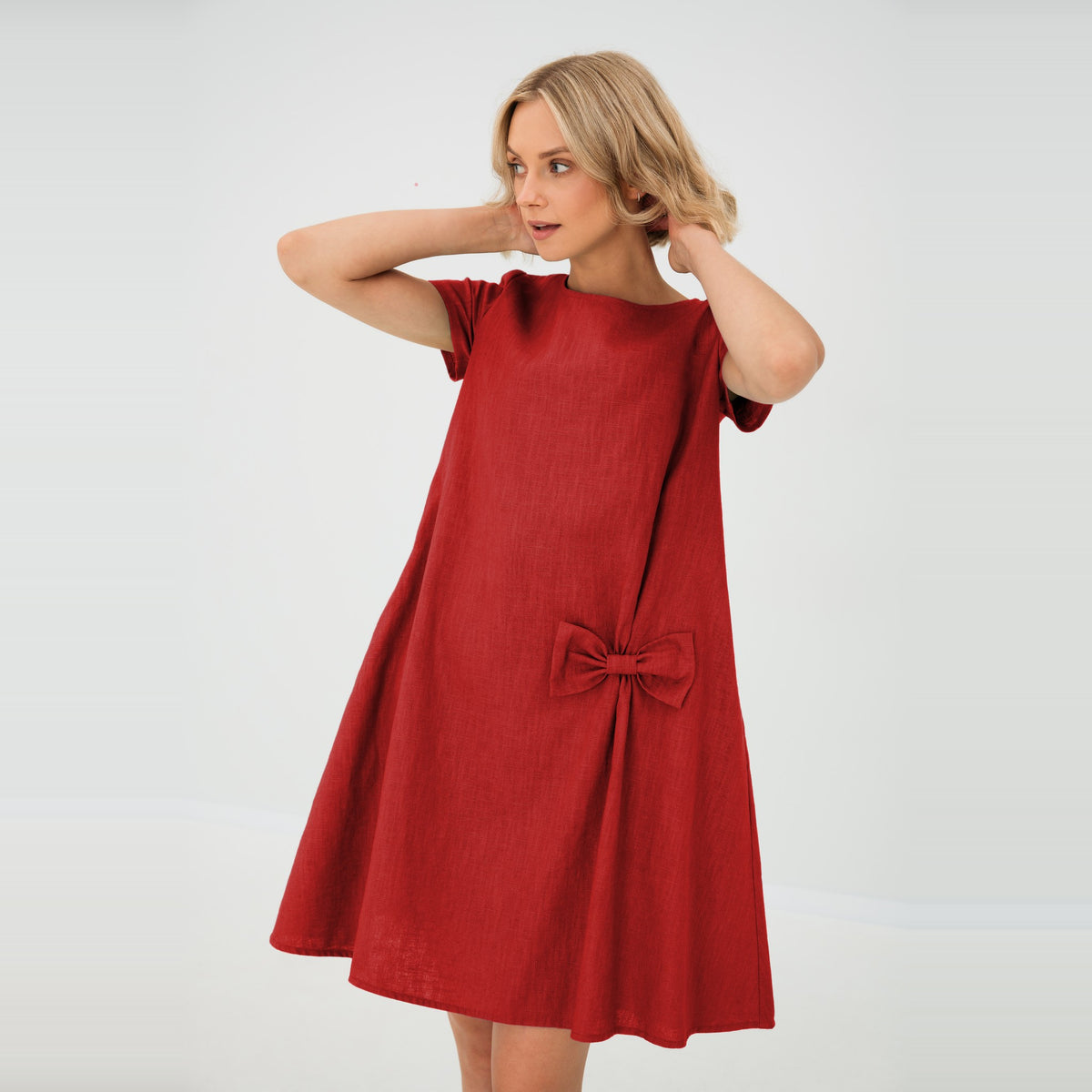 A woman modeling a short-sleeve red linen dress with a gathered bow detail at the front, set against a plain light gray studio background.