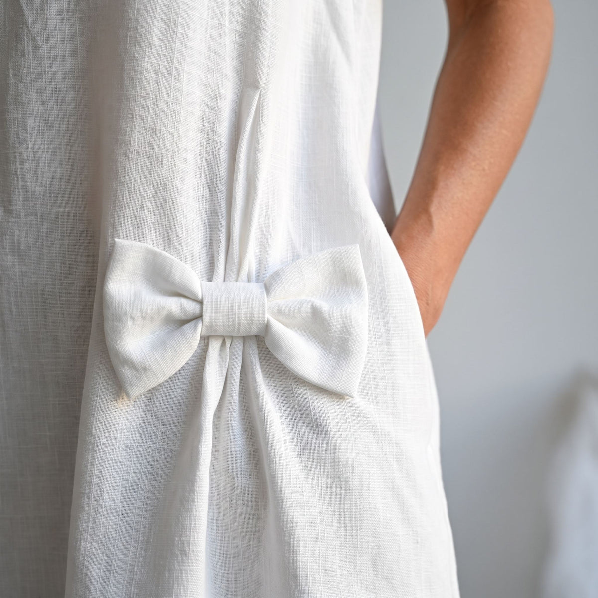 Close-up of a white linen dress with a large fabric bow detail at the waist, worn by a model whose arm is visible against a soft light gray background.