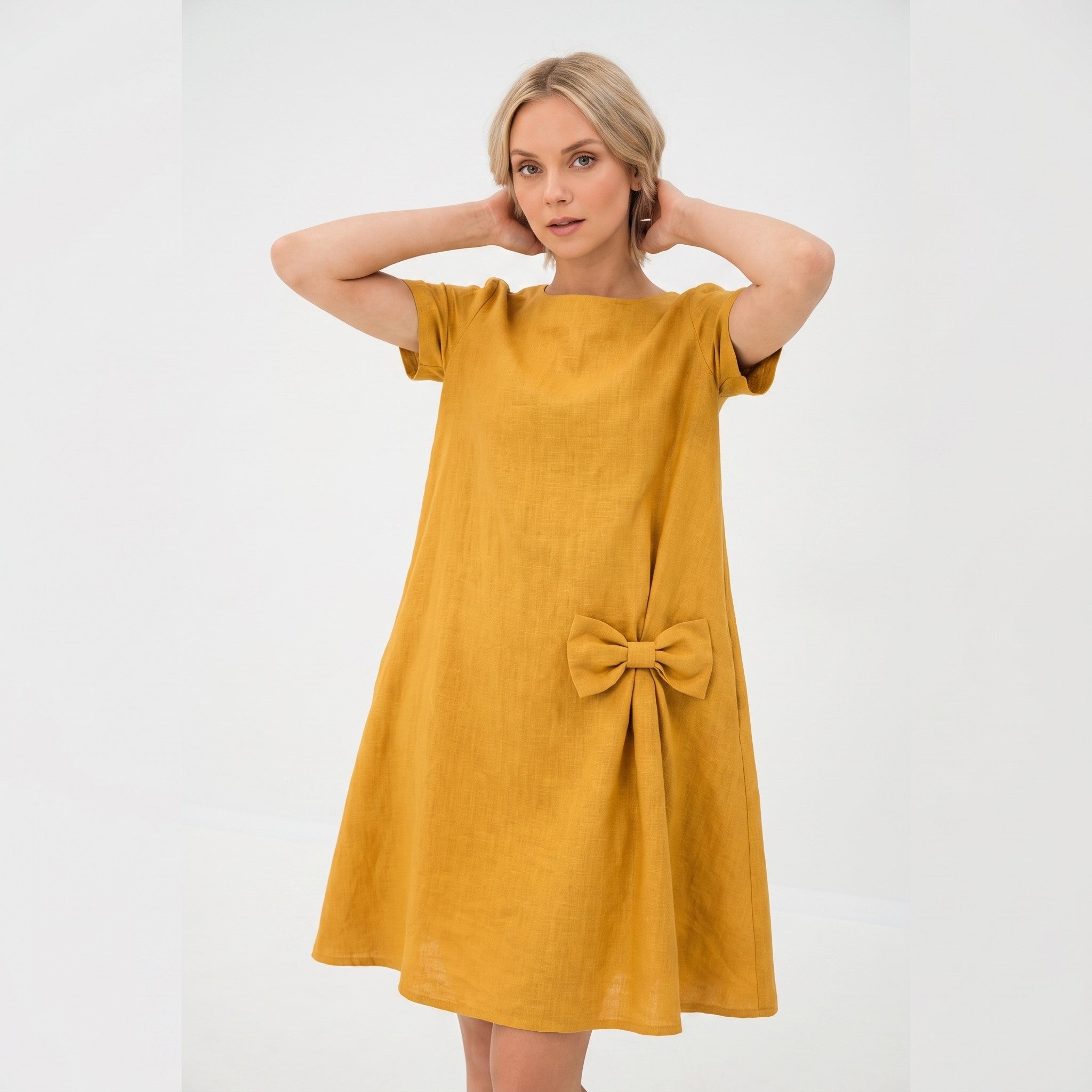 A woman modeling a mustard yellow linen dress with short sleeves and a large bow detail on the front, set against a plain white studio background.