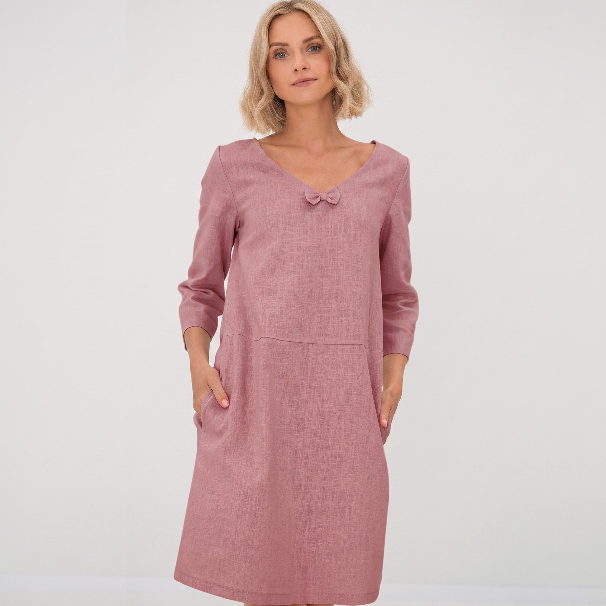 A woman modeling a dusty rose pink long-sleeve linen dress with a small bow detail at the neckline, set against a plain light gray studio background.