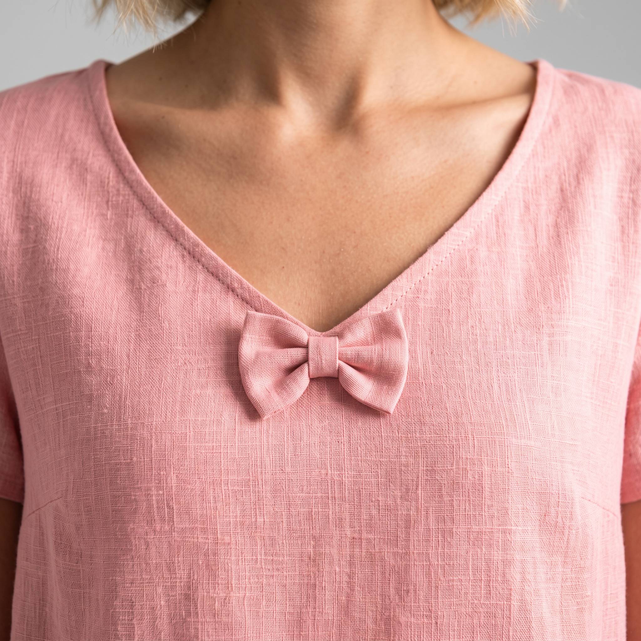 A close-up of a person wearing a light pink linen V-neck top with a matching fabric bow detail at the center of the neckline.