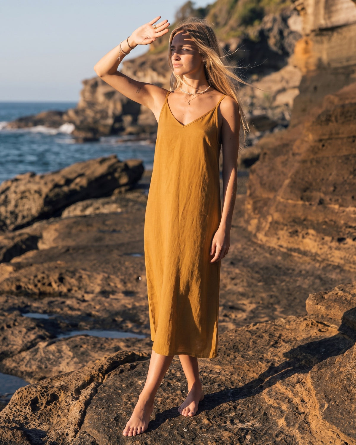 LeMuse SUNSET linen dress in ochre – model in motion with arm raised on the cliffs #Color_Ochre