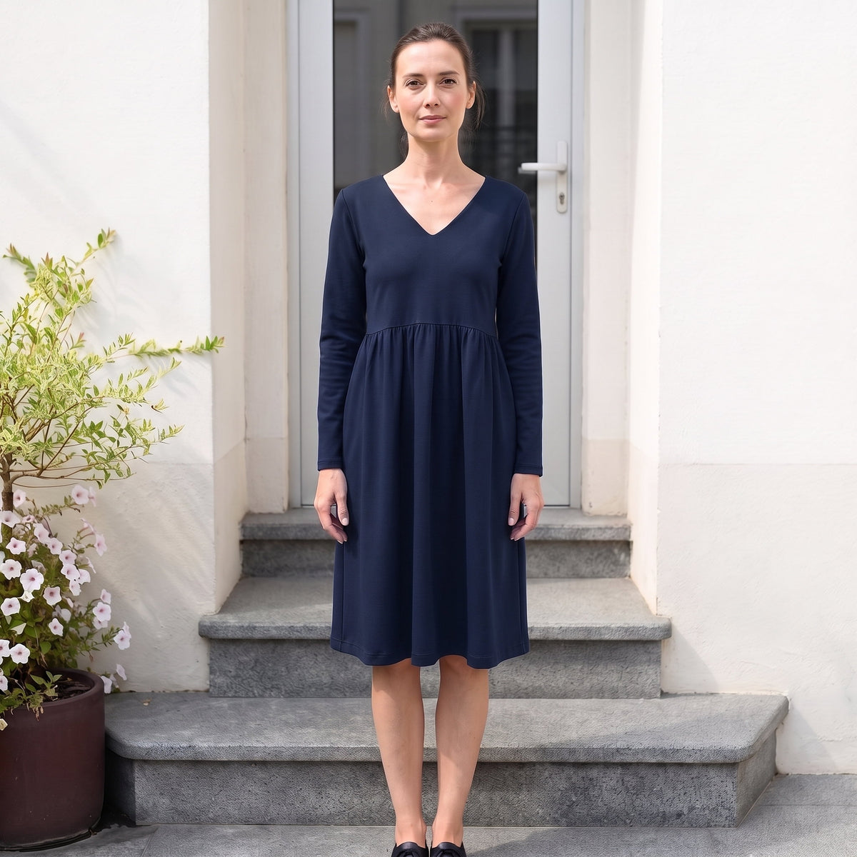 A woman wearing a navy blue long-sleeve midi dress stands on stone steps in front of a white building wall, with a potted plant to the left.