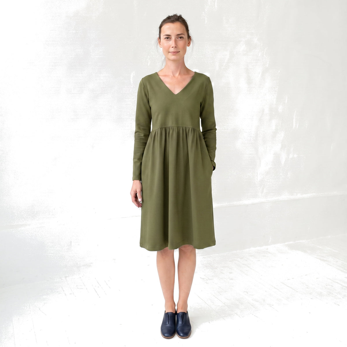 A woman modeling a long-sleeve olive green midi dress with a V-neck and side pockets, standing against a white textured studio wall.