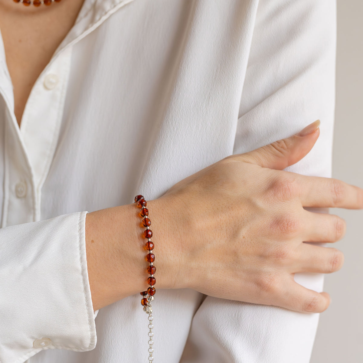A close-up of a person wearing a beaded bracelet on their wrist, dressed in a white button-down shirt. The bracelet features polished reddish-brown beads with a silver-toned metal extender chain.