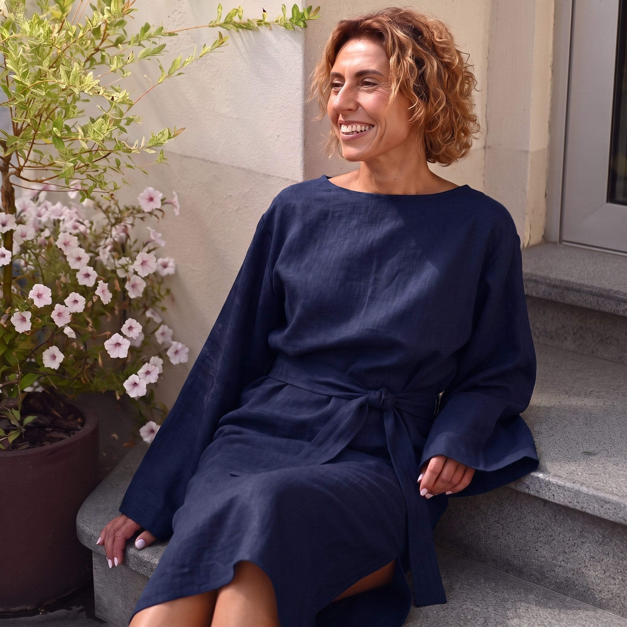 A woman with curly light brown hair sits smiling on outdoor stone steps, wearing a loose navy blue long-sleeve dress with a tie waist. A potted plant with white flowers is visible to her left.