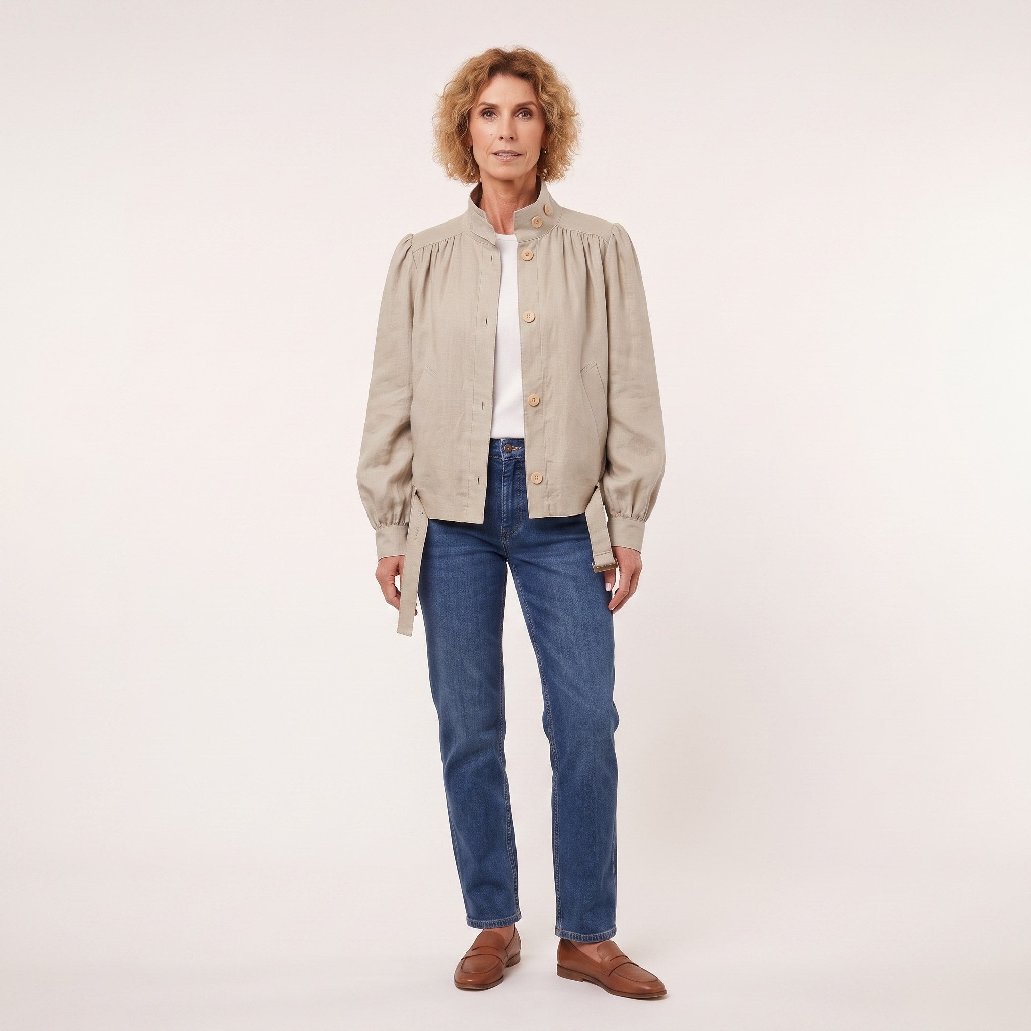 A woman modeling a light beige linen jacket with gathered shoulders and gold buttons, worn over a white top and blue jeans, against a plain off-white studio background.